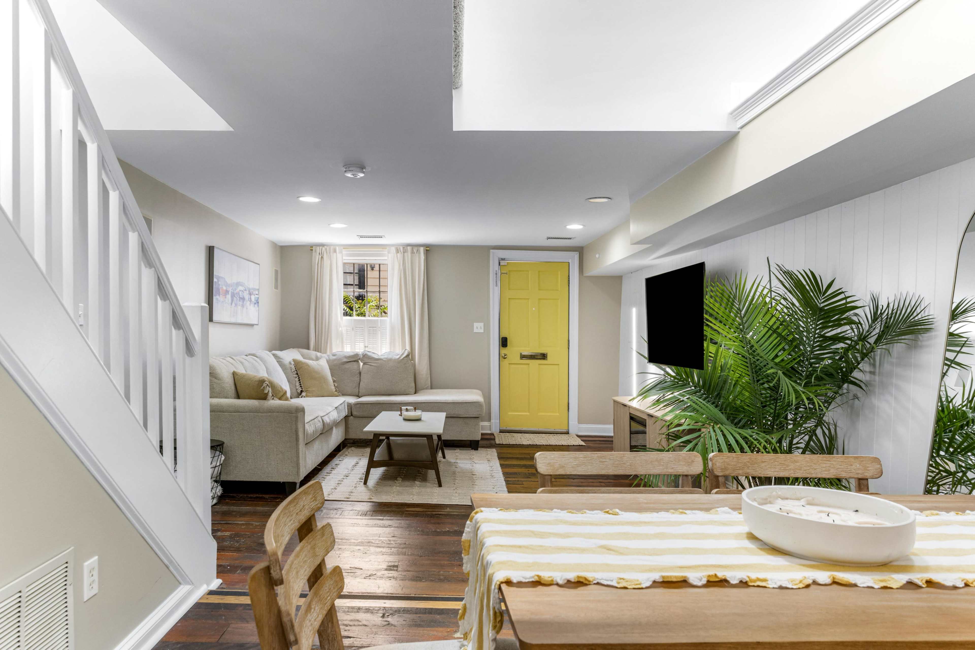 The image shows a cozy, well-lit living space with a beige sofa, a wooden dining table, and a yellow front door, along with decorative plants.