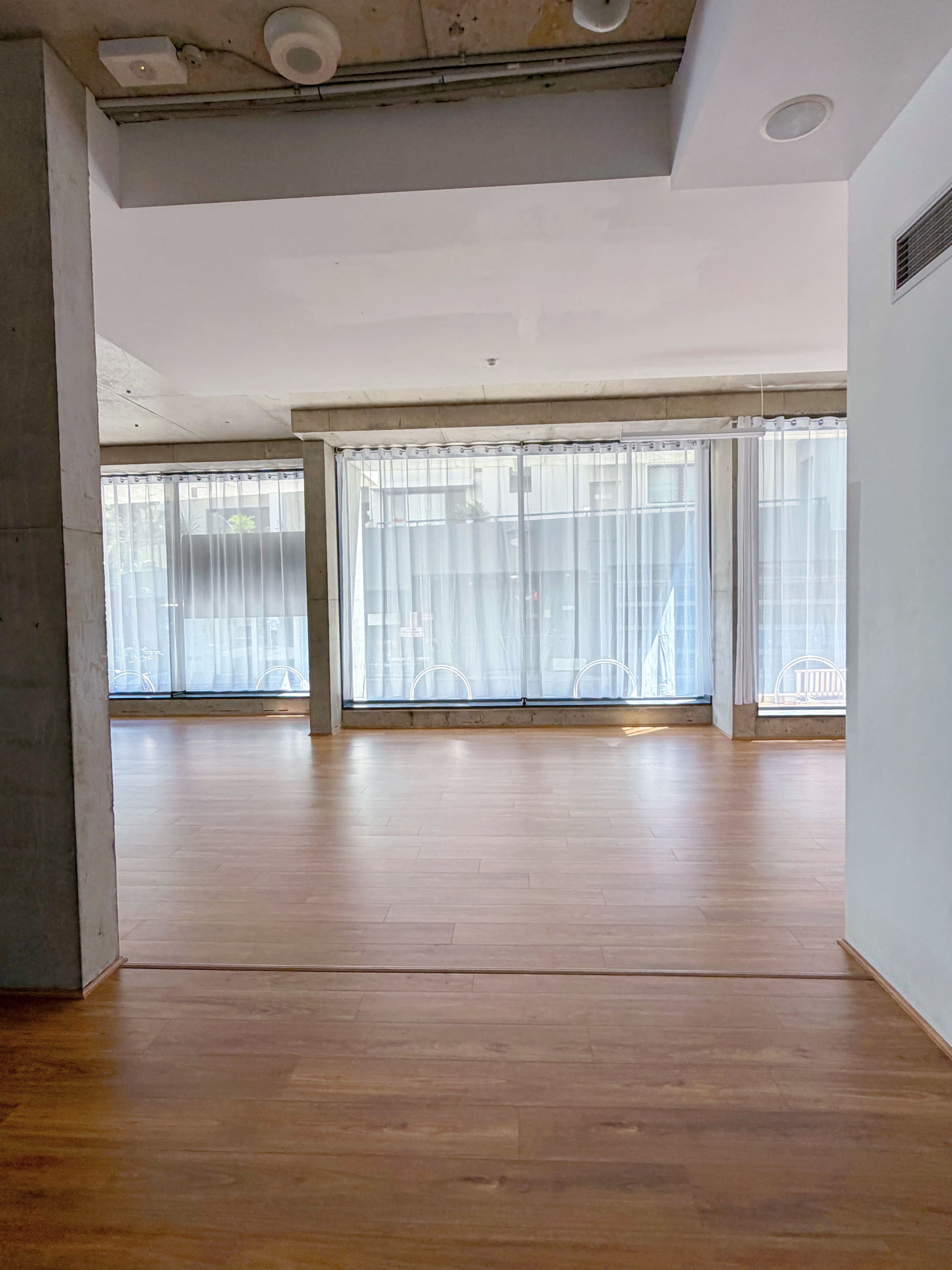 The image shows an empty room with wooden flooring and large windows that allow natural light to enter.
