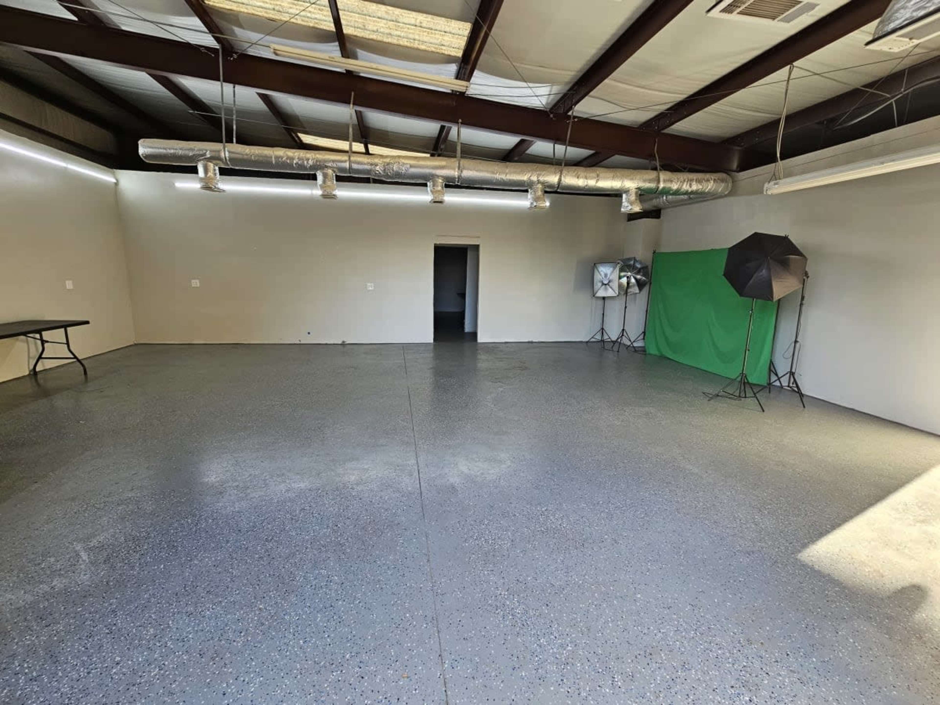 A spacious, empty room with a concrete floor, a folding table, a green backdrop, and studio lighting setup in the corners.