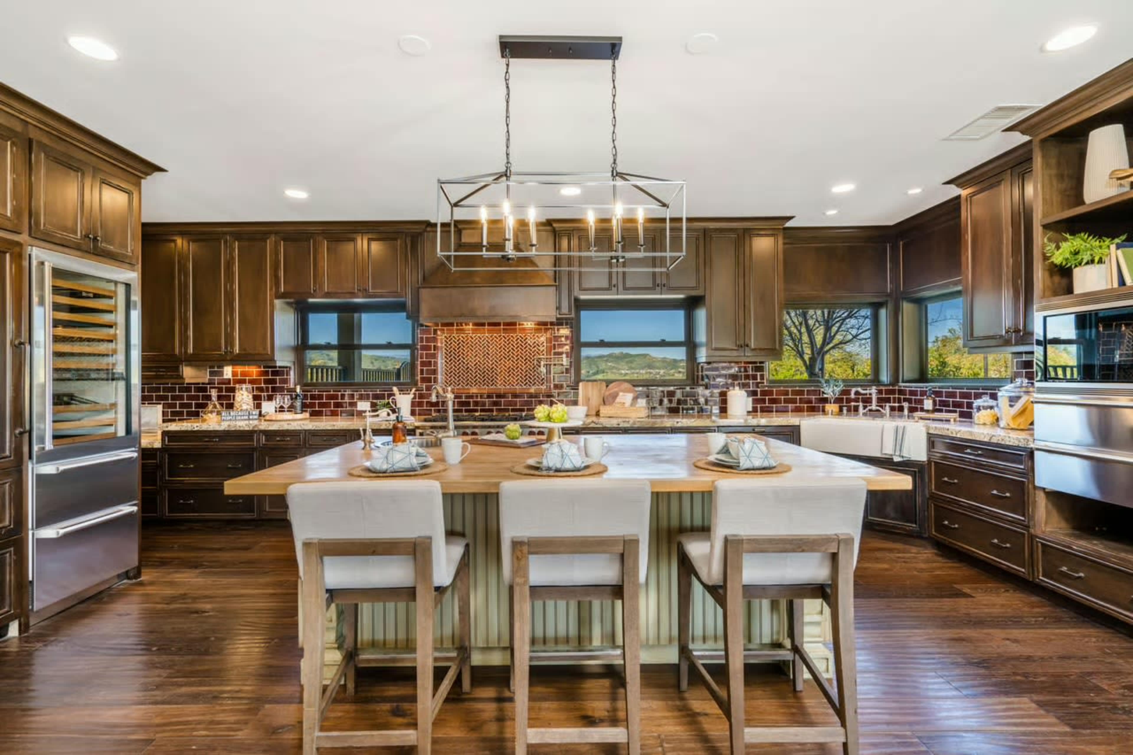 The image shows a modern kitchen with dark wood cabinetry, a large island with seating, and stainless steel appliances, highlighted by natural light from large windows.
