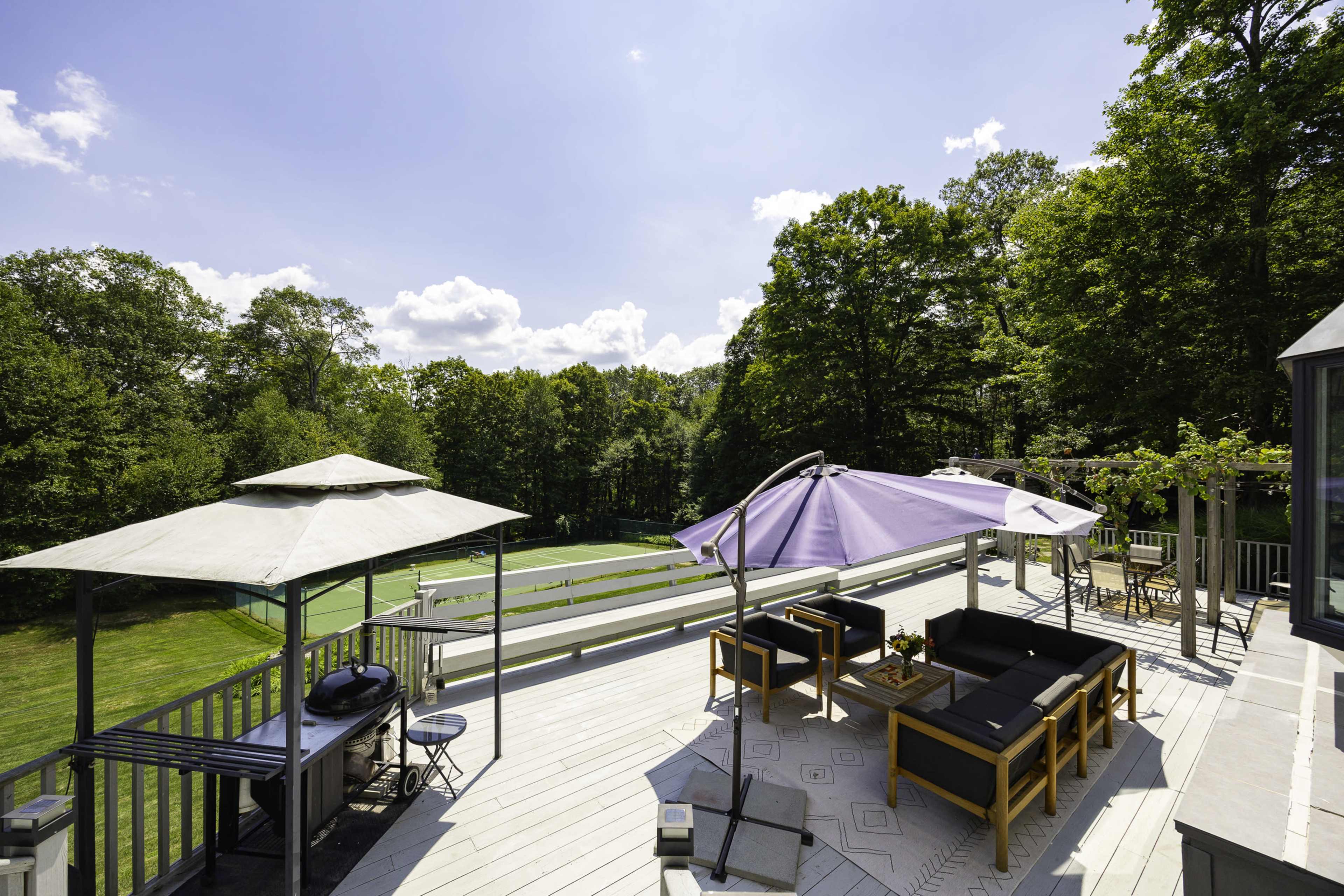 A spacious deck features seating areas shaded by umbrellas, overlooking a green lawn and tennis court surrounded by trees.