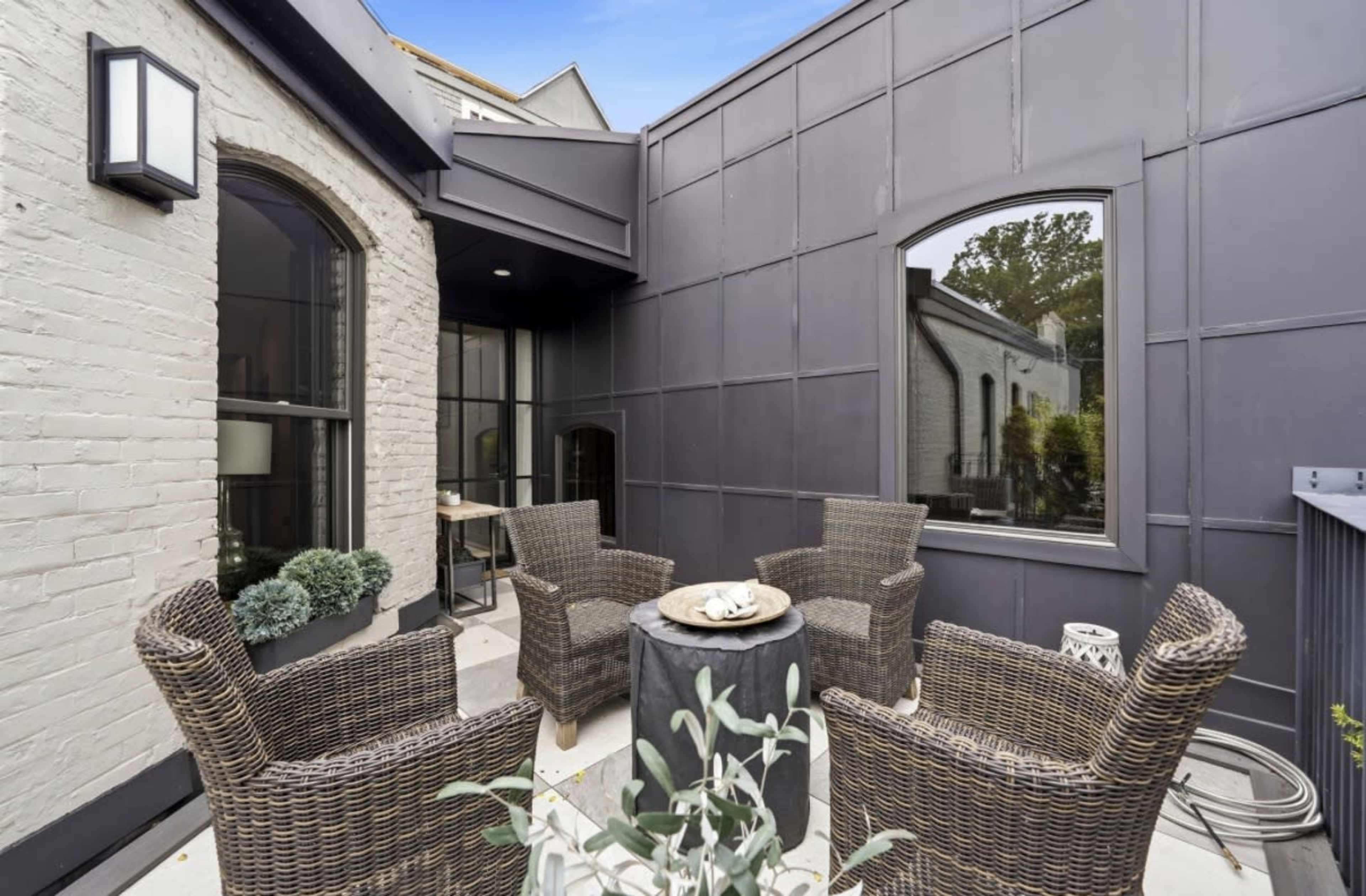 The image shows a small outdoor patio area with four wicker chairs arranged around a round table, framed by dark wall panels and large windows.