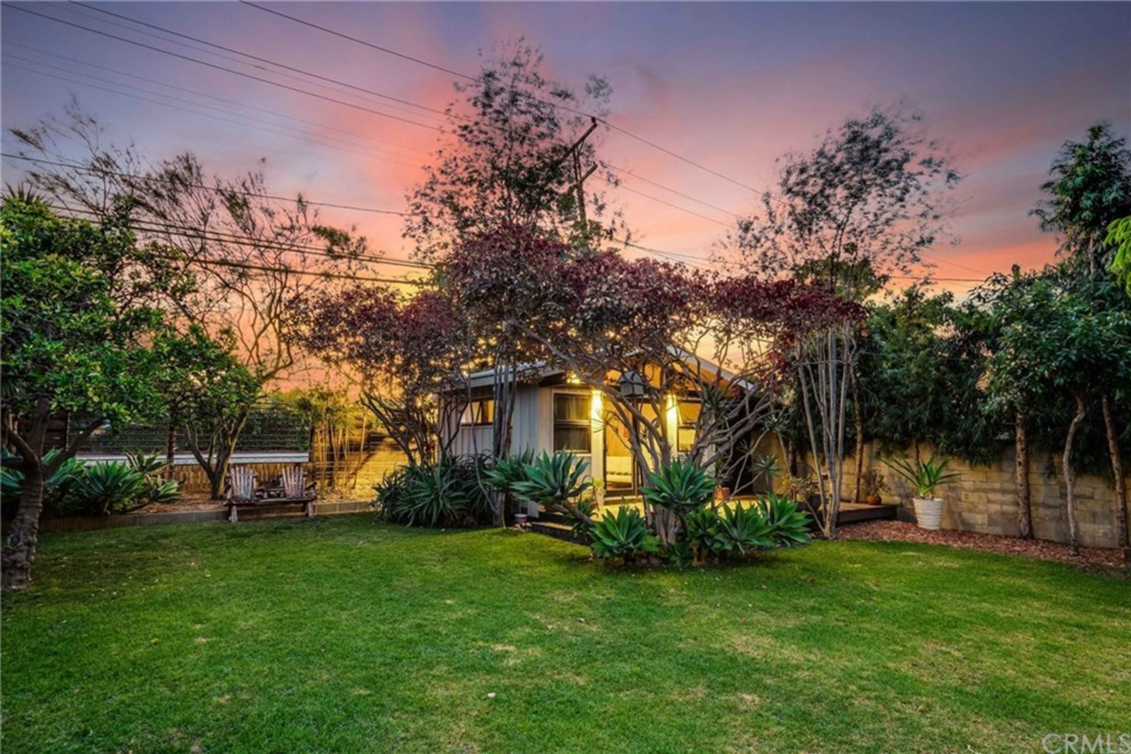 A small house is set in a lush backyard surrounded by trees and plants, with the sky displaying hues of pink and orange at sunset.