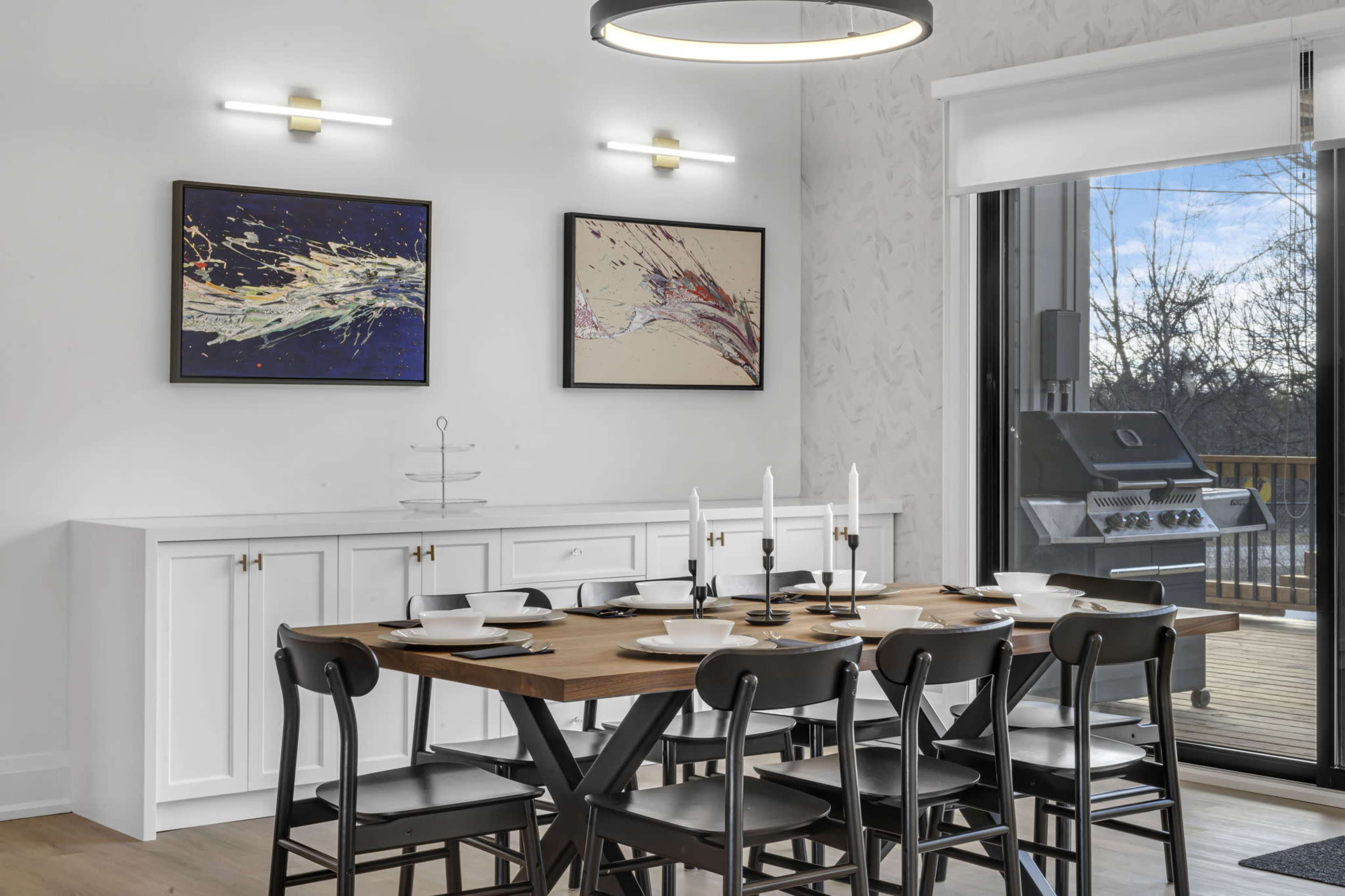 A modern dining area features a wooden table set for meals, black chairs, and two abstract paintings on the walls beside a sliding glass door leading to an outdoor deck with a barbecue grill.