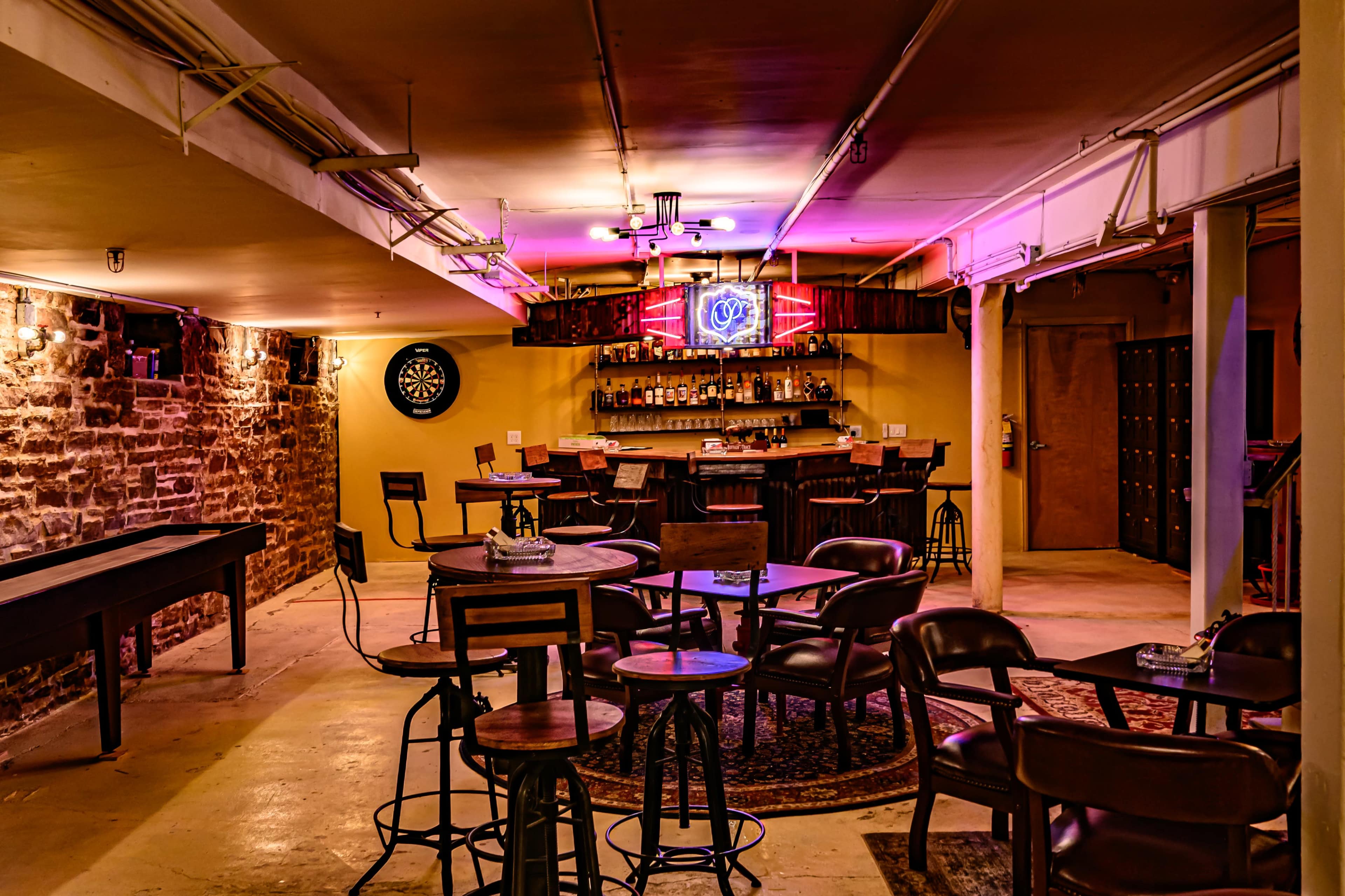 The image shows a dimly lit basement bar featuring a bar counter, several tables, and a dartboard on one wall.