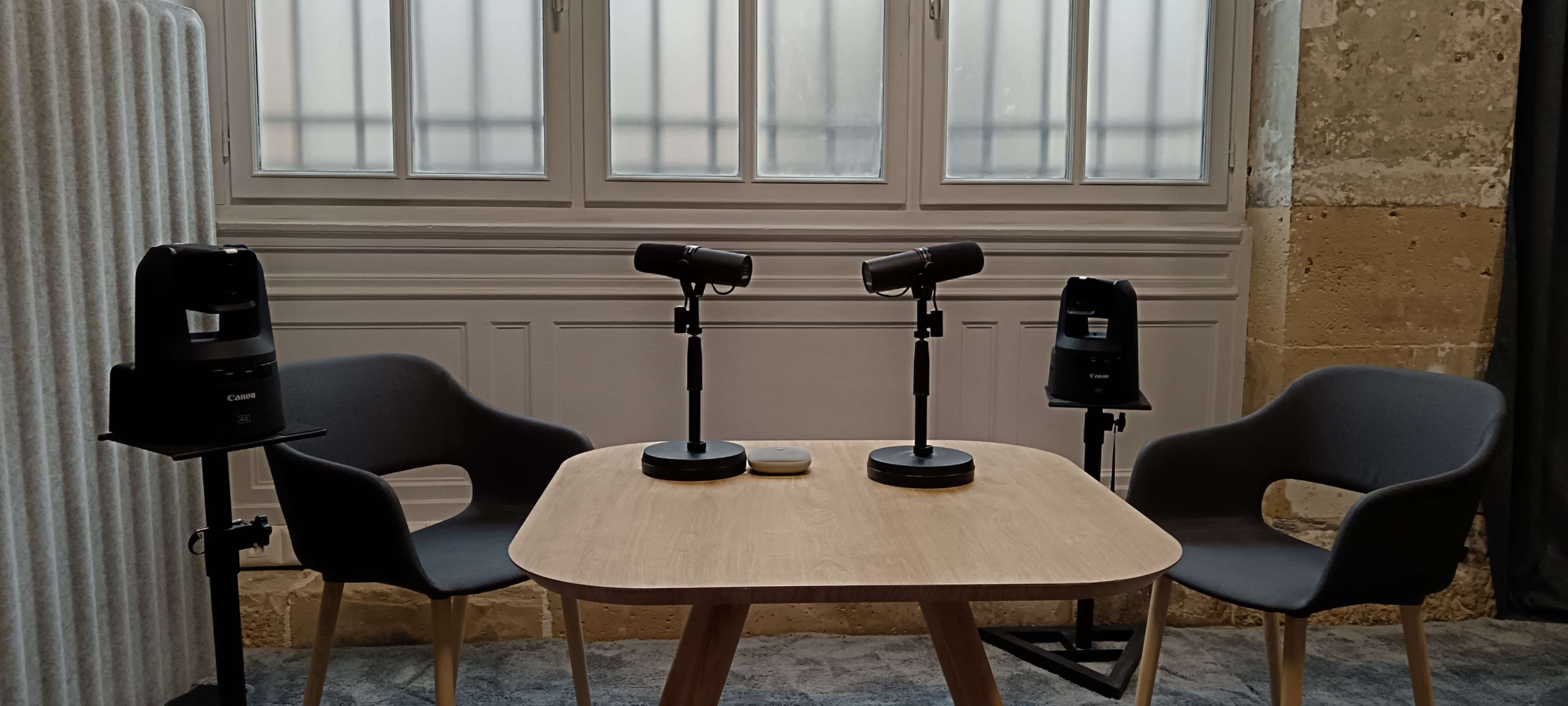 The image shows a minimalist recording setup with two microphones on stands facing a wooden table, flanked by two gray chairs, in a room with large windows.
