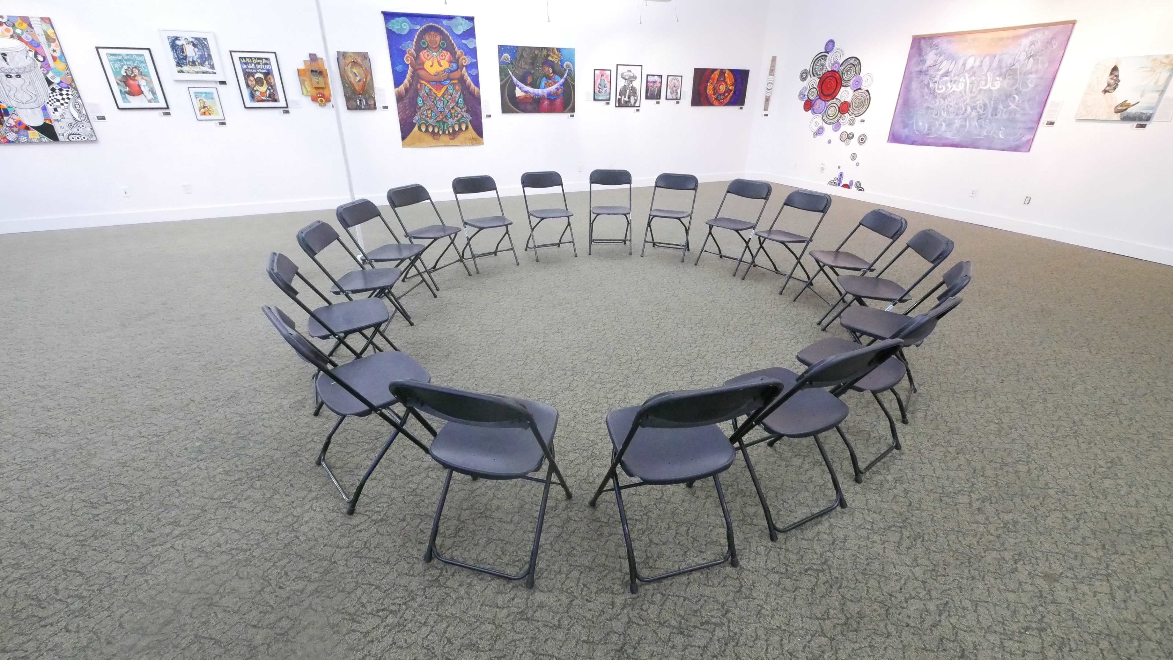 A circular arrangement of twenty black folding chairs in an art gallery with colorful artwork displayed on the walls.