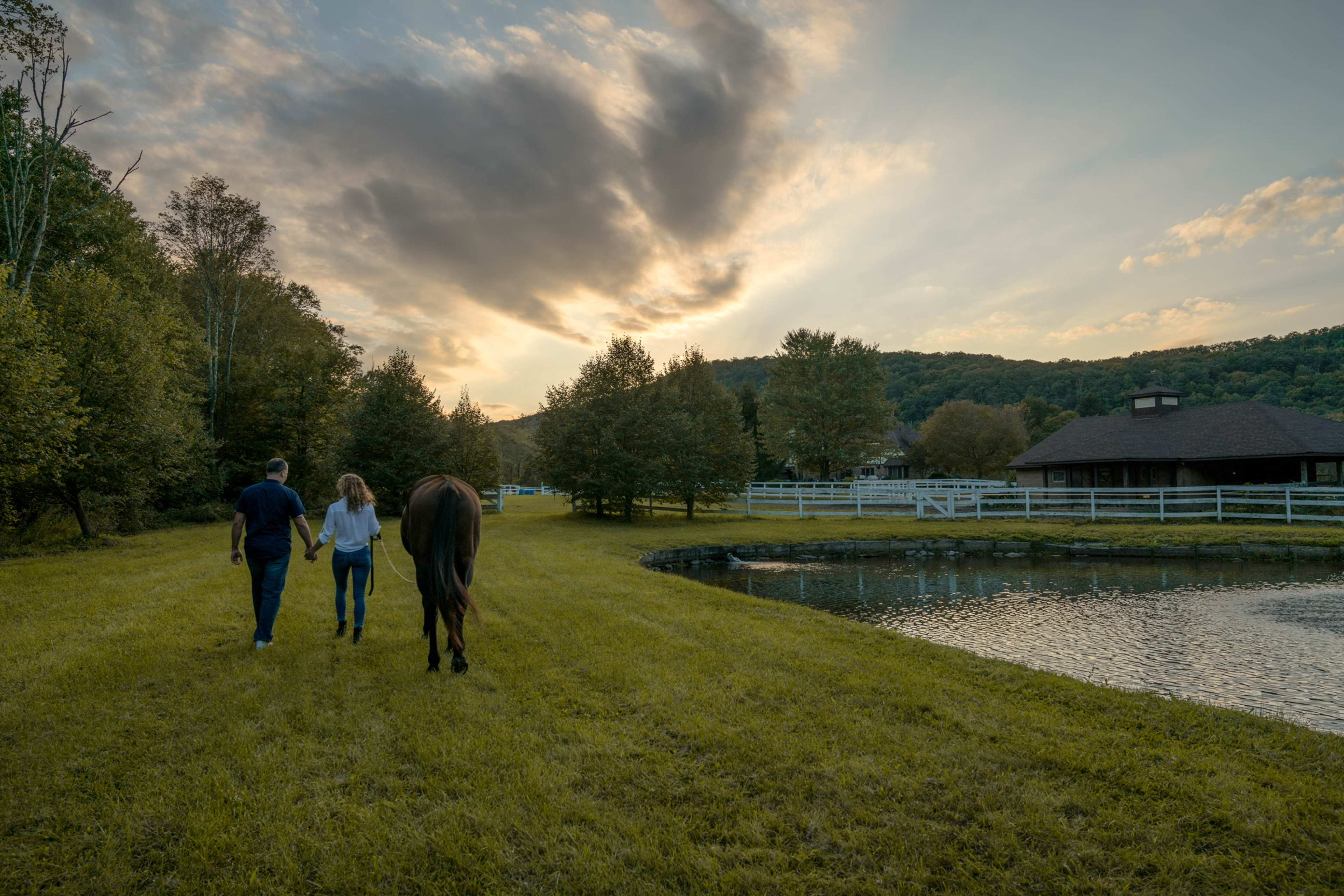 A man and a woman walk alongside a horse on a grassy path near a pond, with trees and a building in the background under a sunset sky.