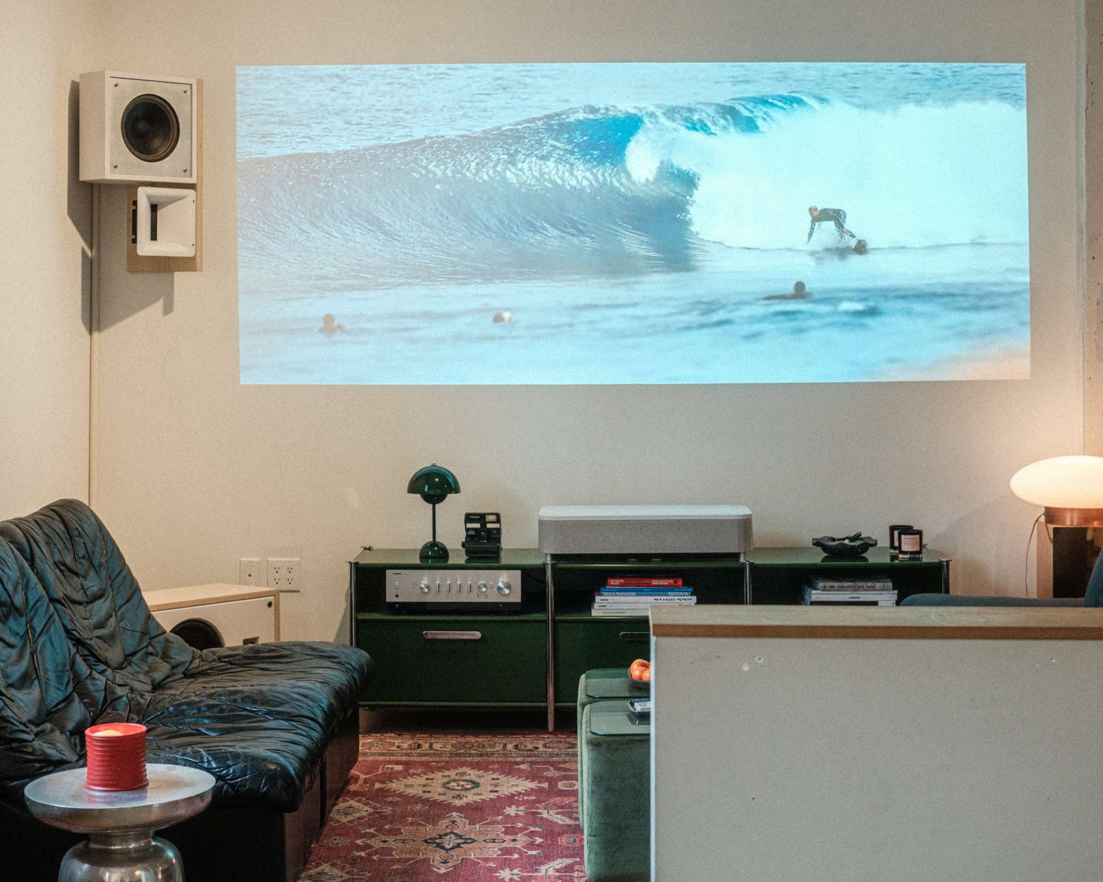 A living room features a projection of a surfer riding a wave on one wall, with a sofa, coffee table, and green storage units nearby.