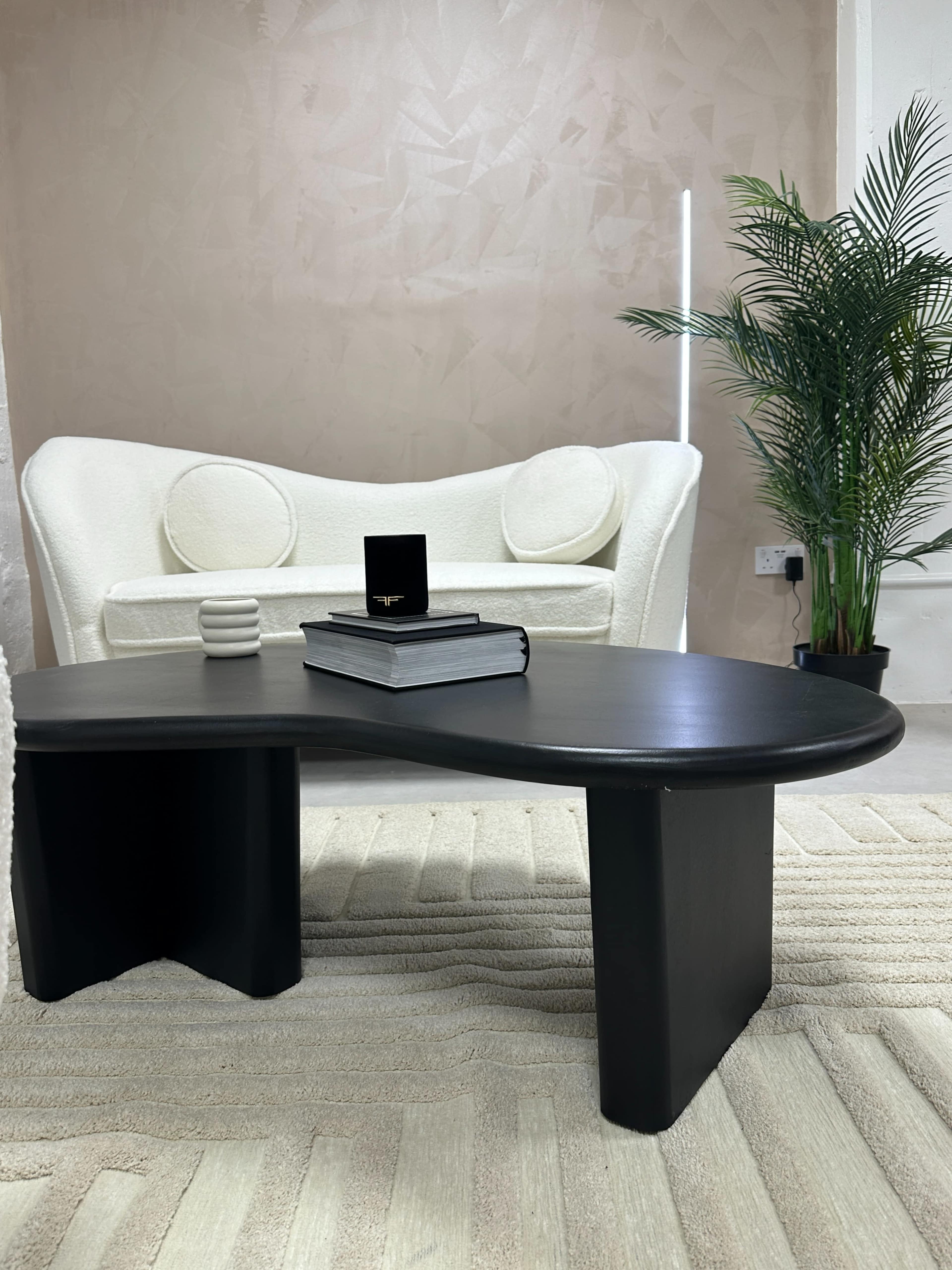 A black, curvy coffee table sits in front of a white upholstered sofa, with a potted plant and a stack of books on top.