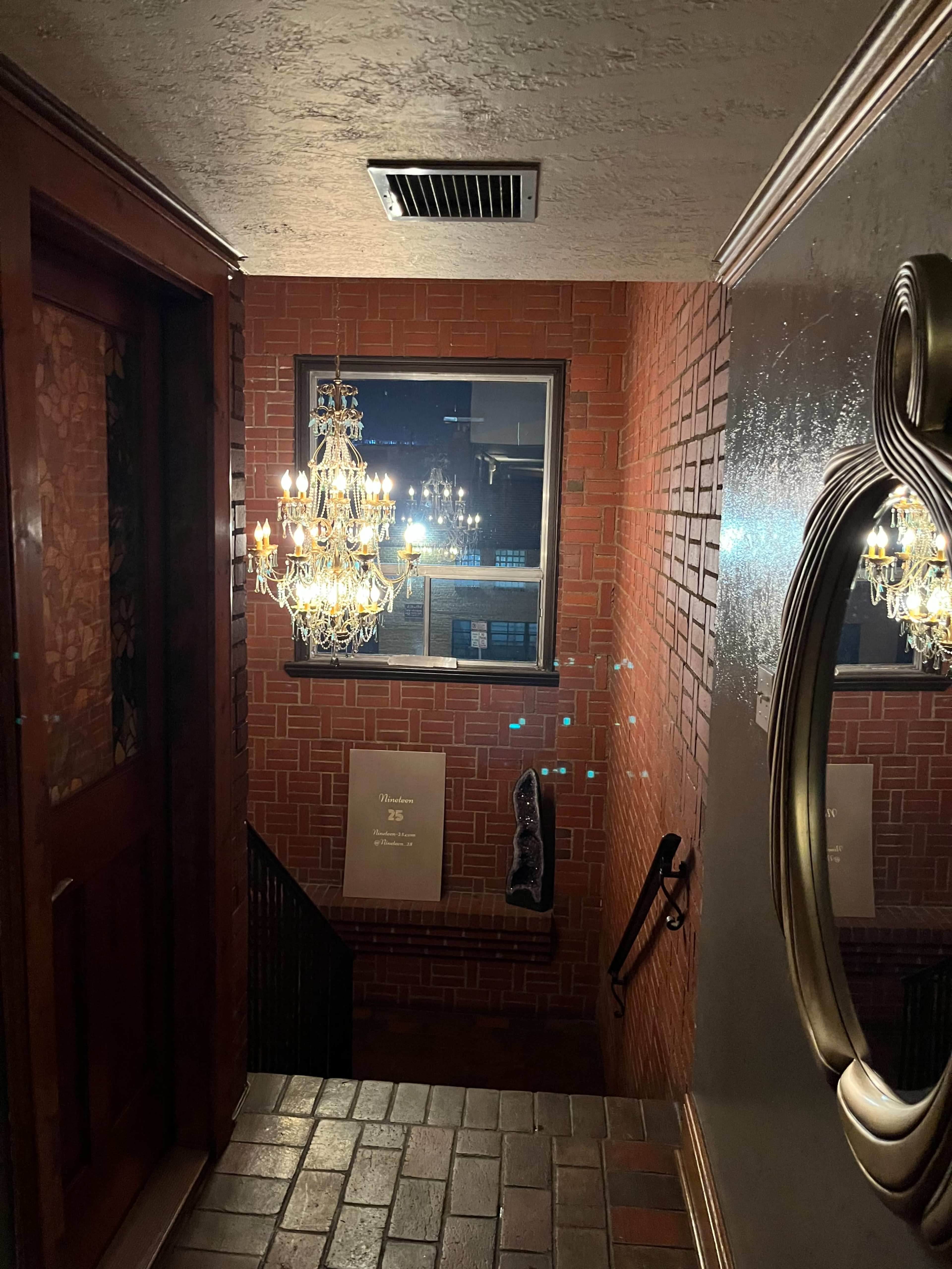 A dimly lit staircase leading downwards, with a large mirror and a chandelier illuminating the area, alongside brick walls and a door at the top.