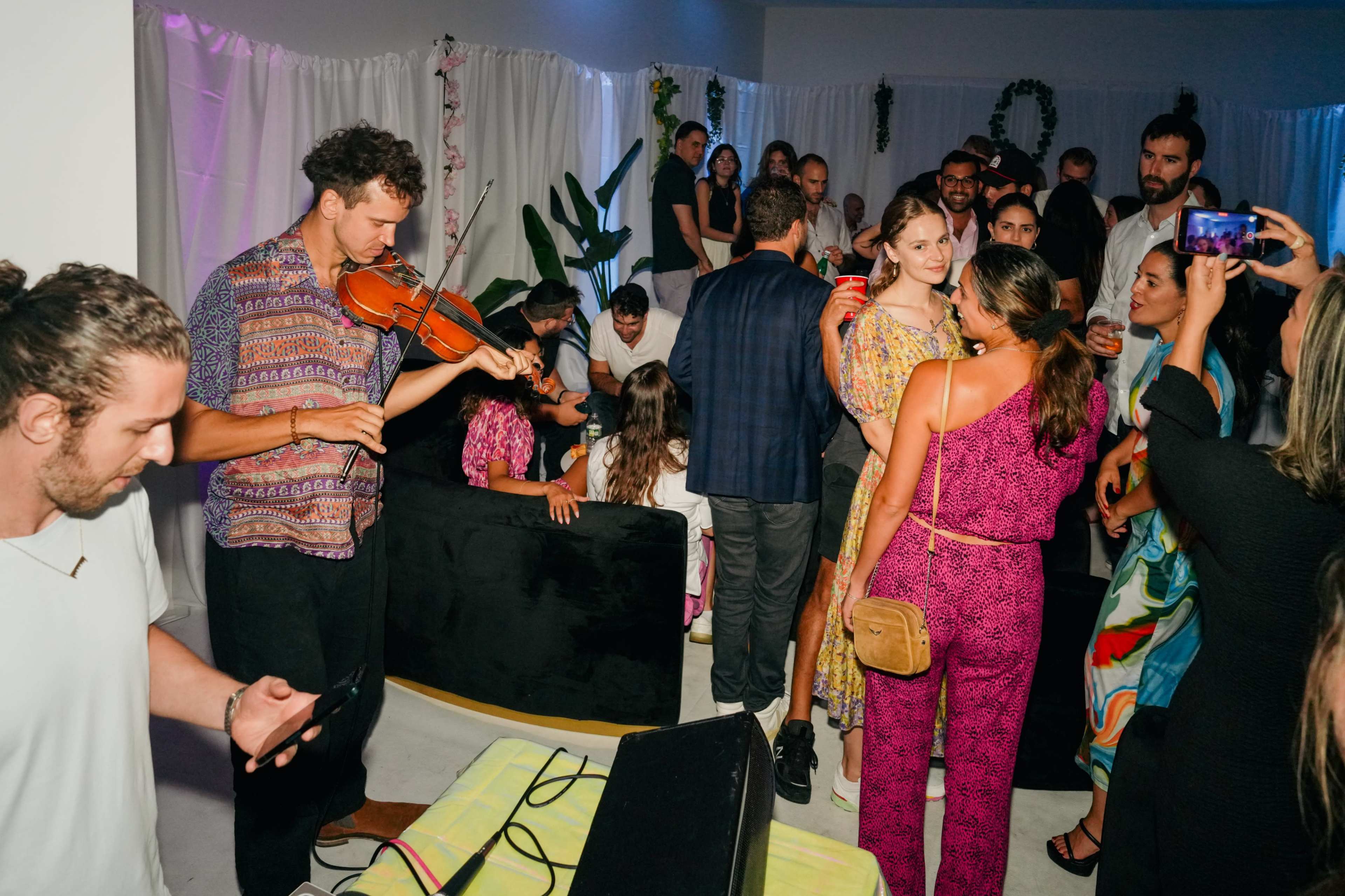A group of people socialize at a vibrant indoor gathering, with one person playing the violin while others chat and take photos.