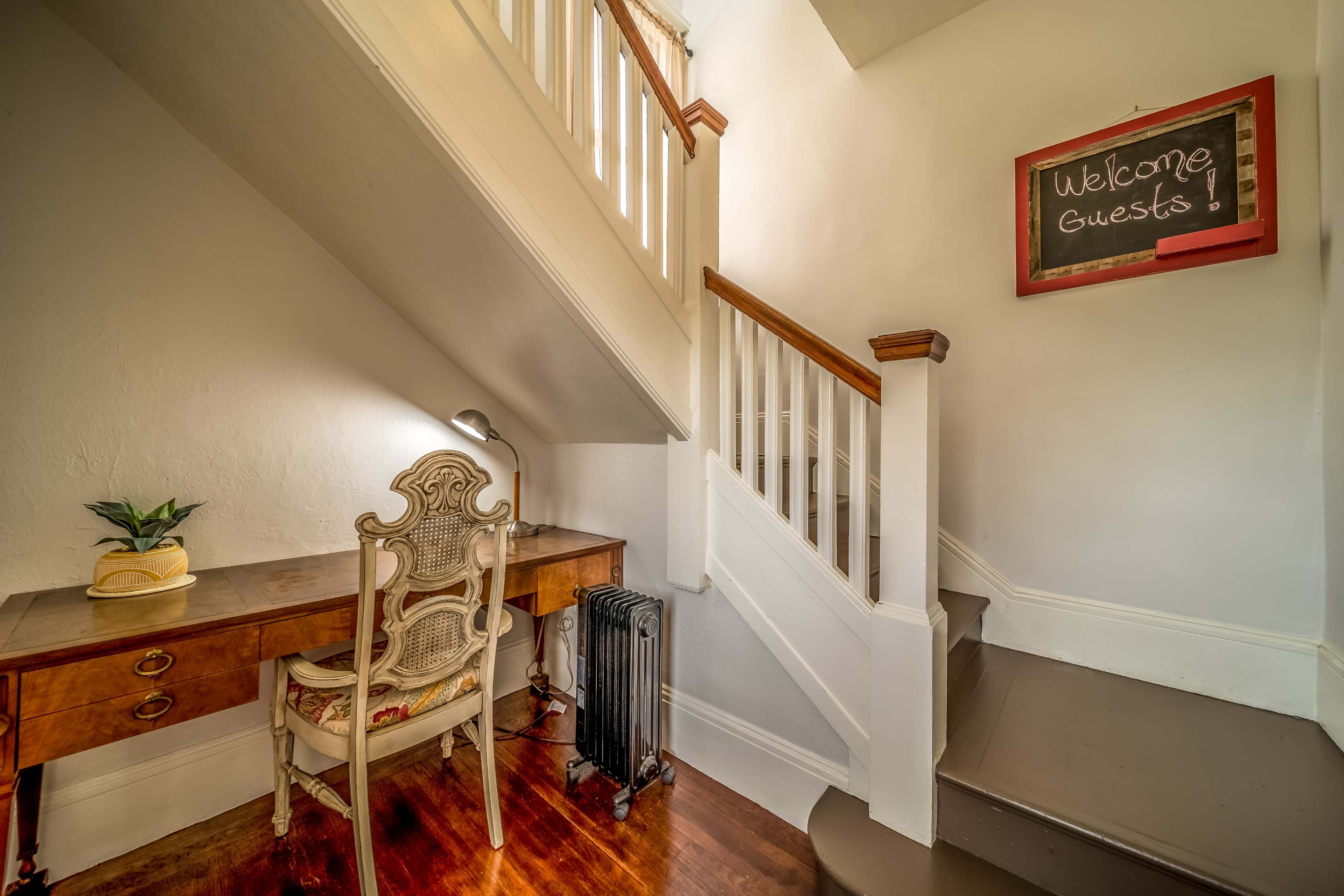 A small entryway features a wooden desk with a chair, a lamp, and a chalkboard that reads "Welcome Guests," next to a staircase.