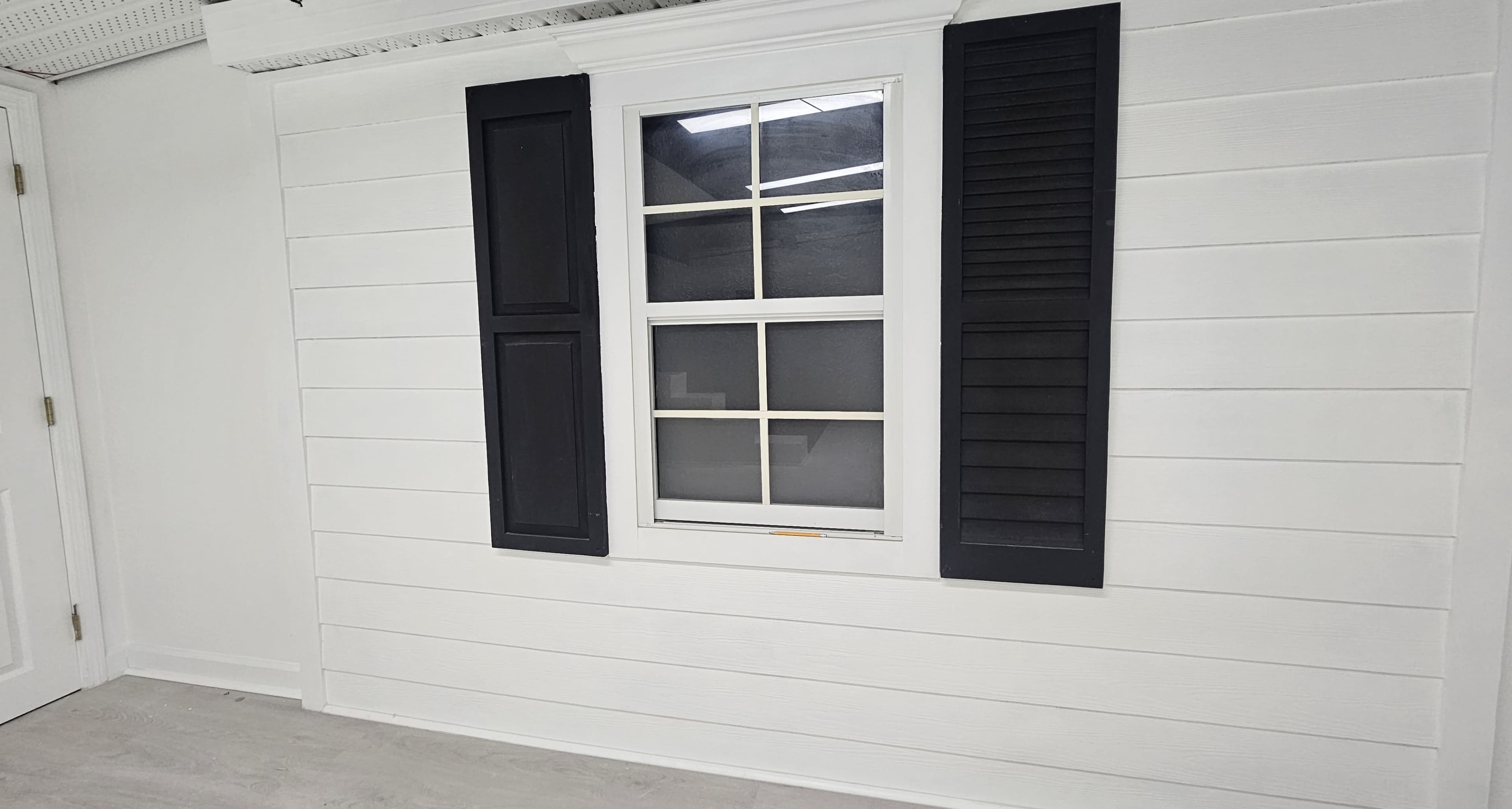 The image shows a white wall with a window featuring black shutters on either side.