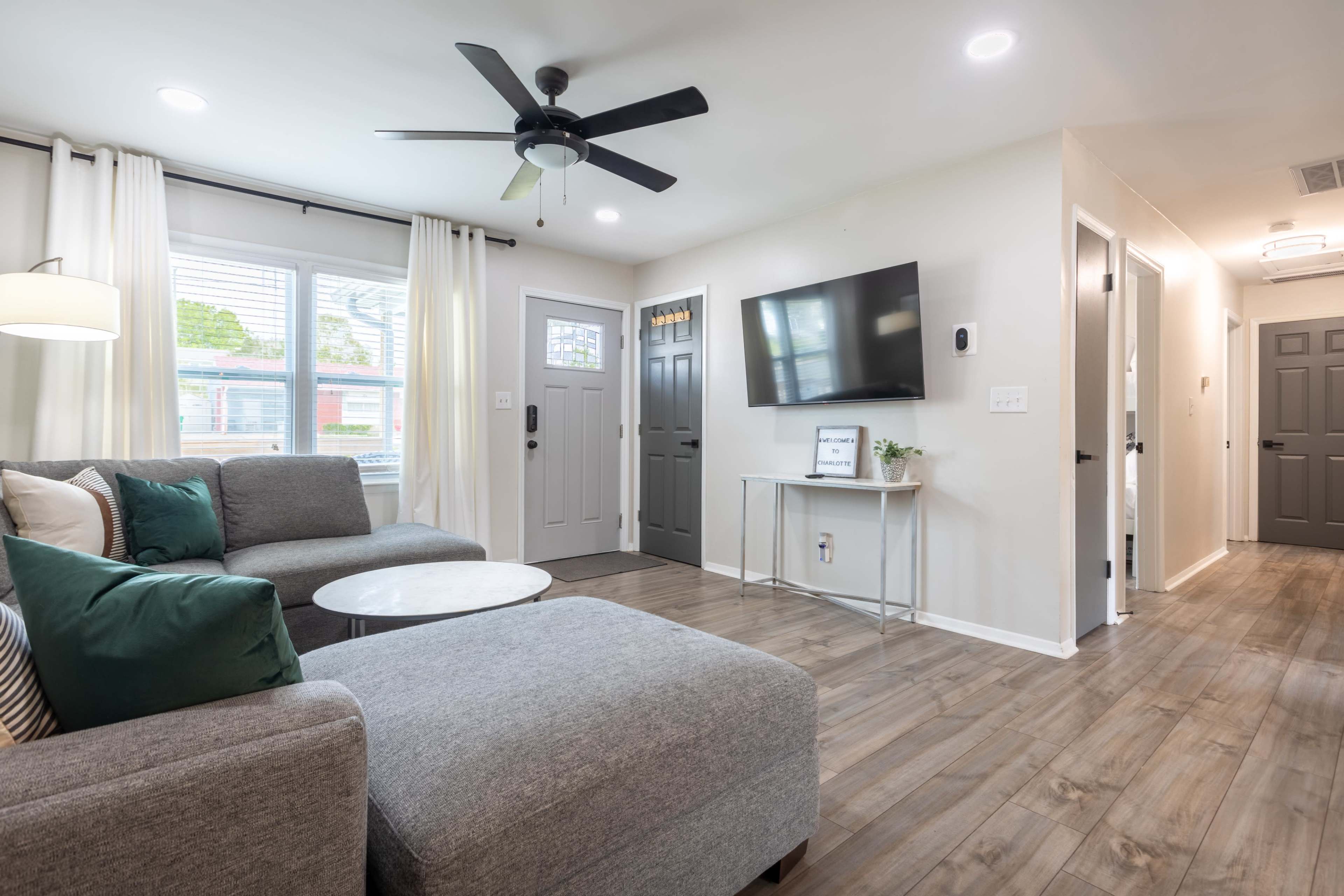 A modern living room featuring a gray sectional sofa, a circular coffee table, a ceiling fan, and a mounted television on a light-colored wall.