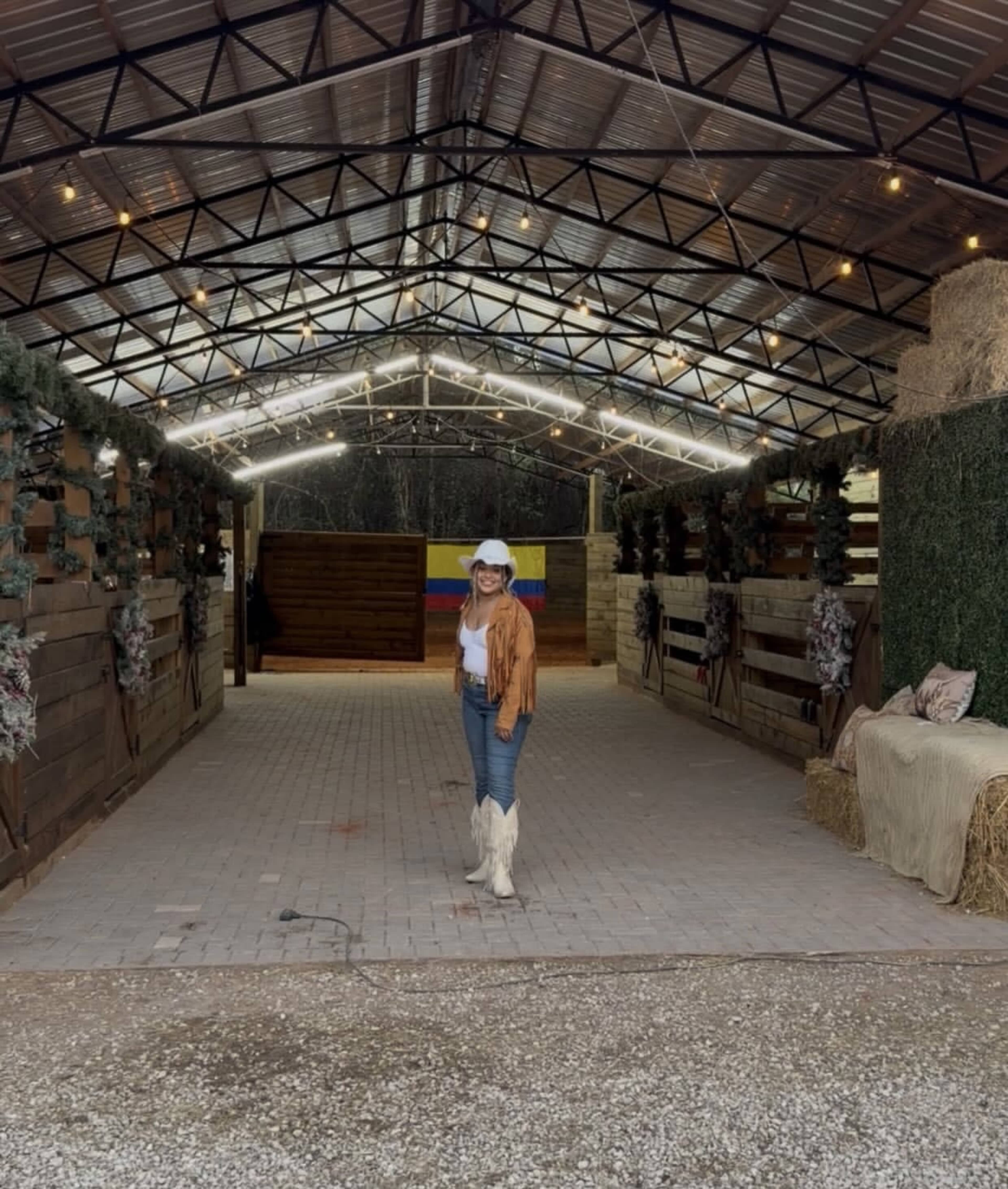 A woman in a white hat and brown cardigan stands in an open barn with dim lighting and decorative greenery.