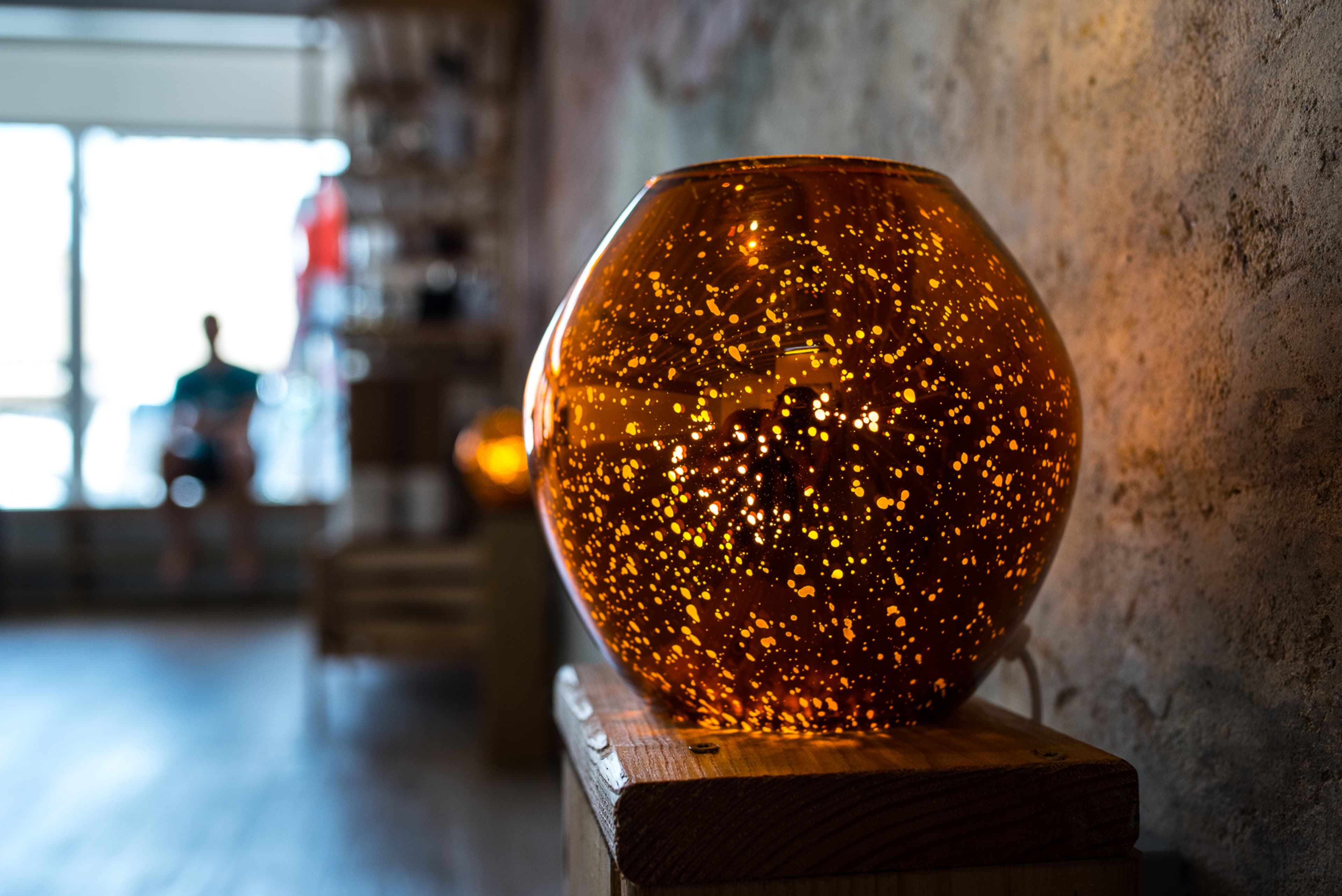 A glowing amber glass lamp with speckles sits on a wooden stand in a warmly lit room with a window visible in the background.