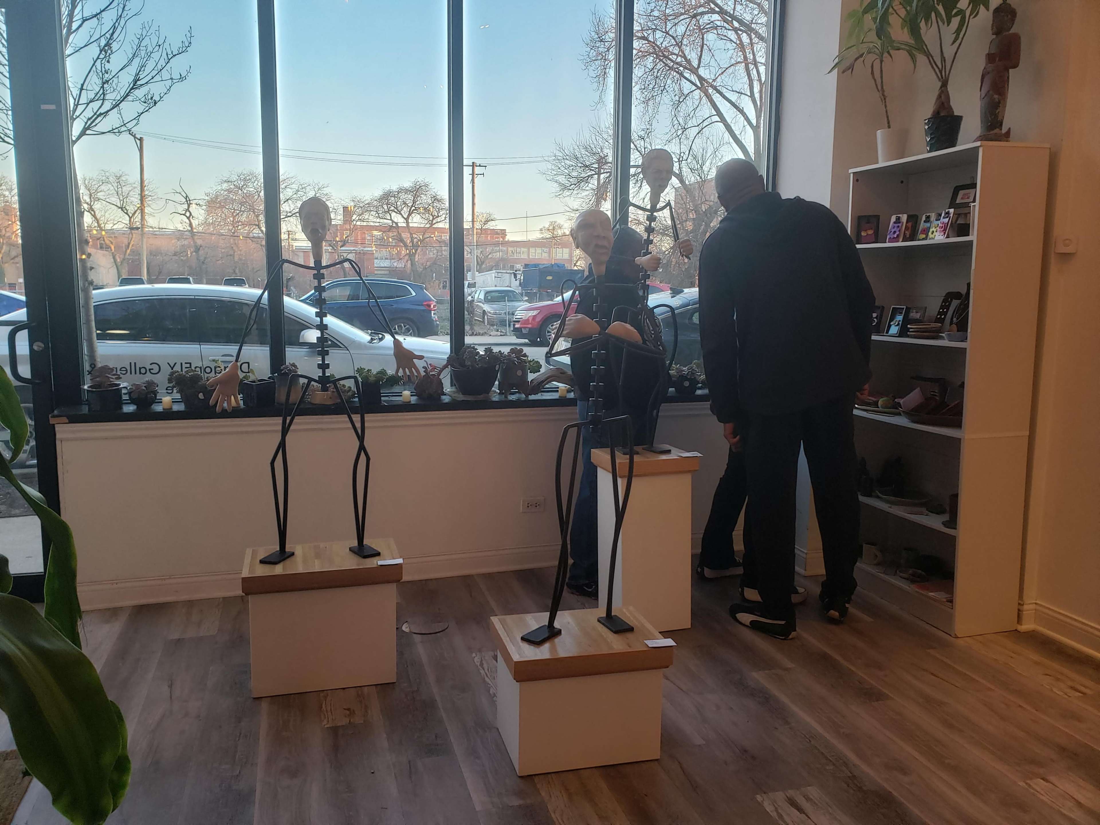 Two mannequins are displayed on pedestals in a shop with large windows, while two customers examine items on a shelf nearby.
