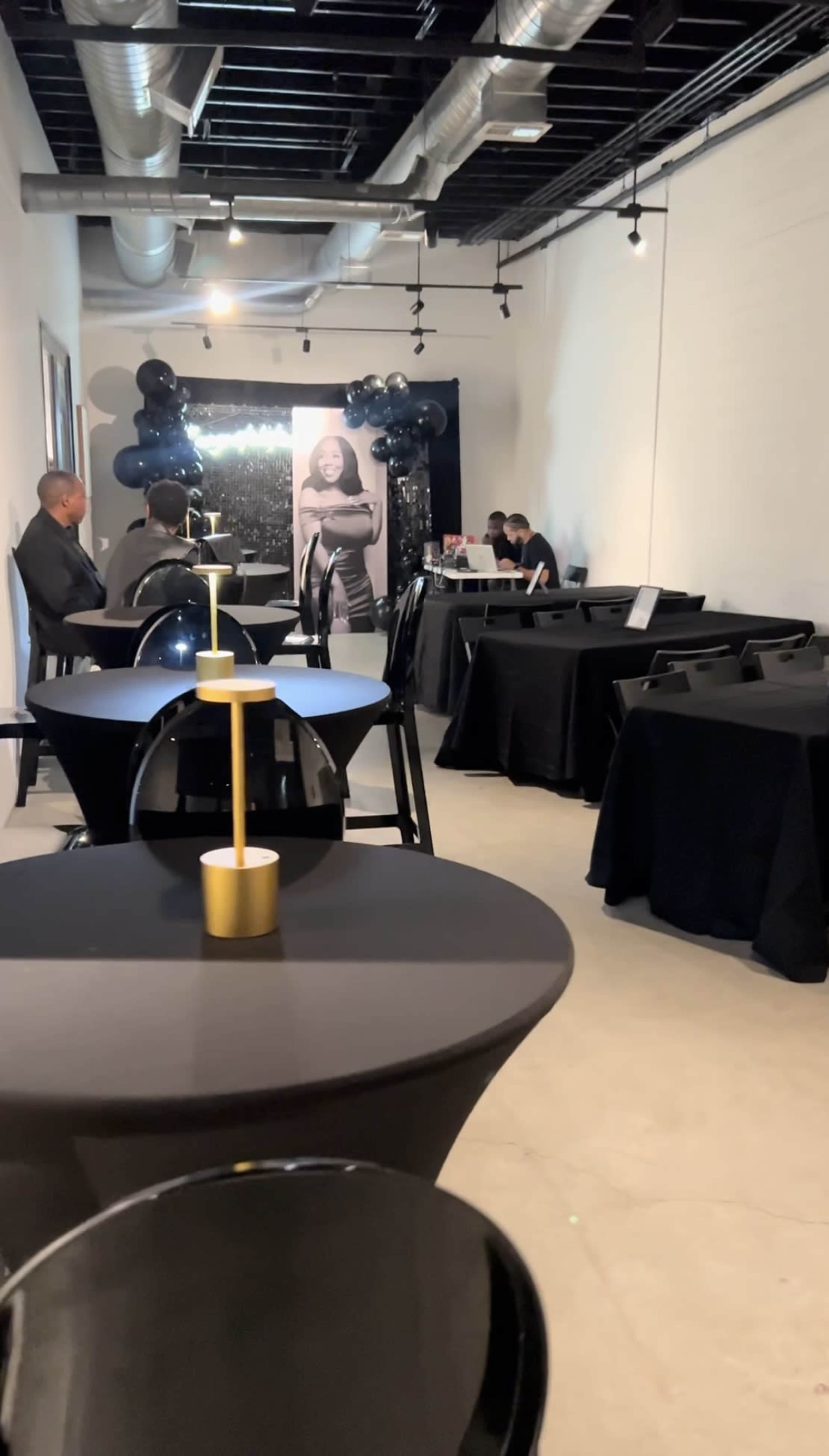 A modern event space is set up with black tables and decor, featuring a backdrop of black balloons and a large portrait.