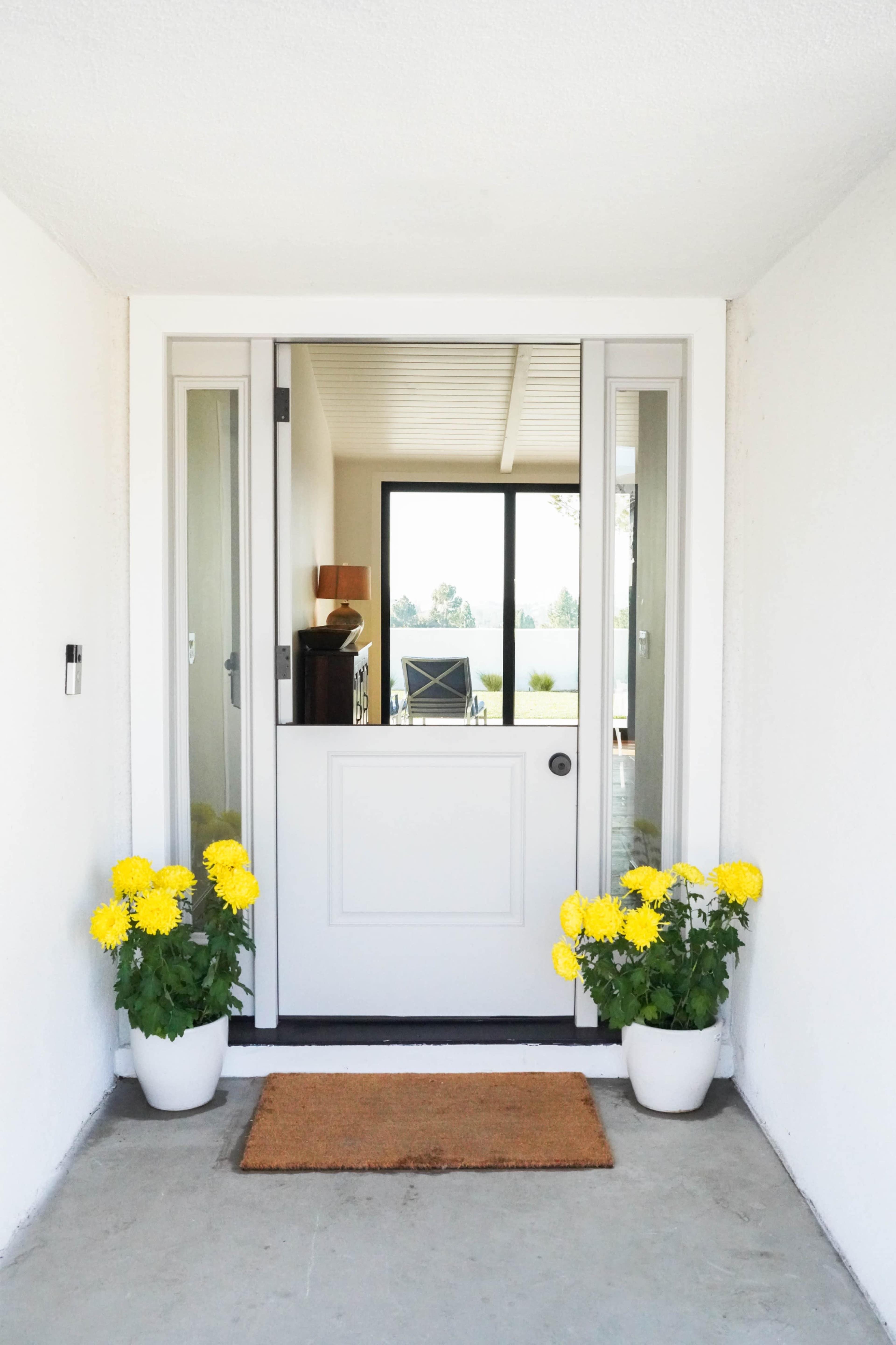 A front entrance with a white door framed by glass panels and two pots of yellow flowers on either side of a welcome mat.