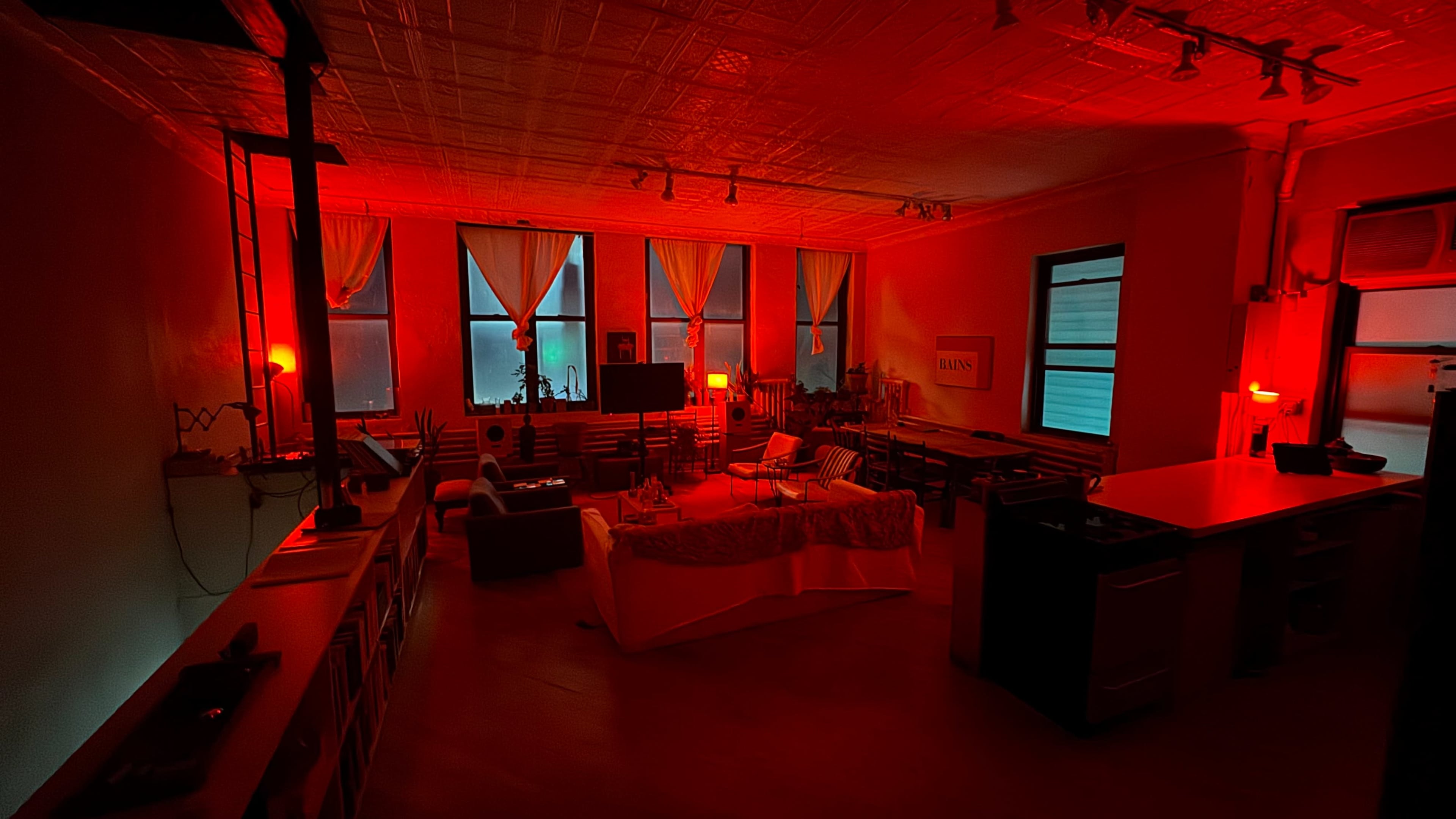The room is illuminated with red lighting, featuring furniture arranged around a central table and windows covered by sheer curtains.