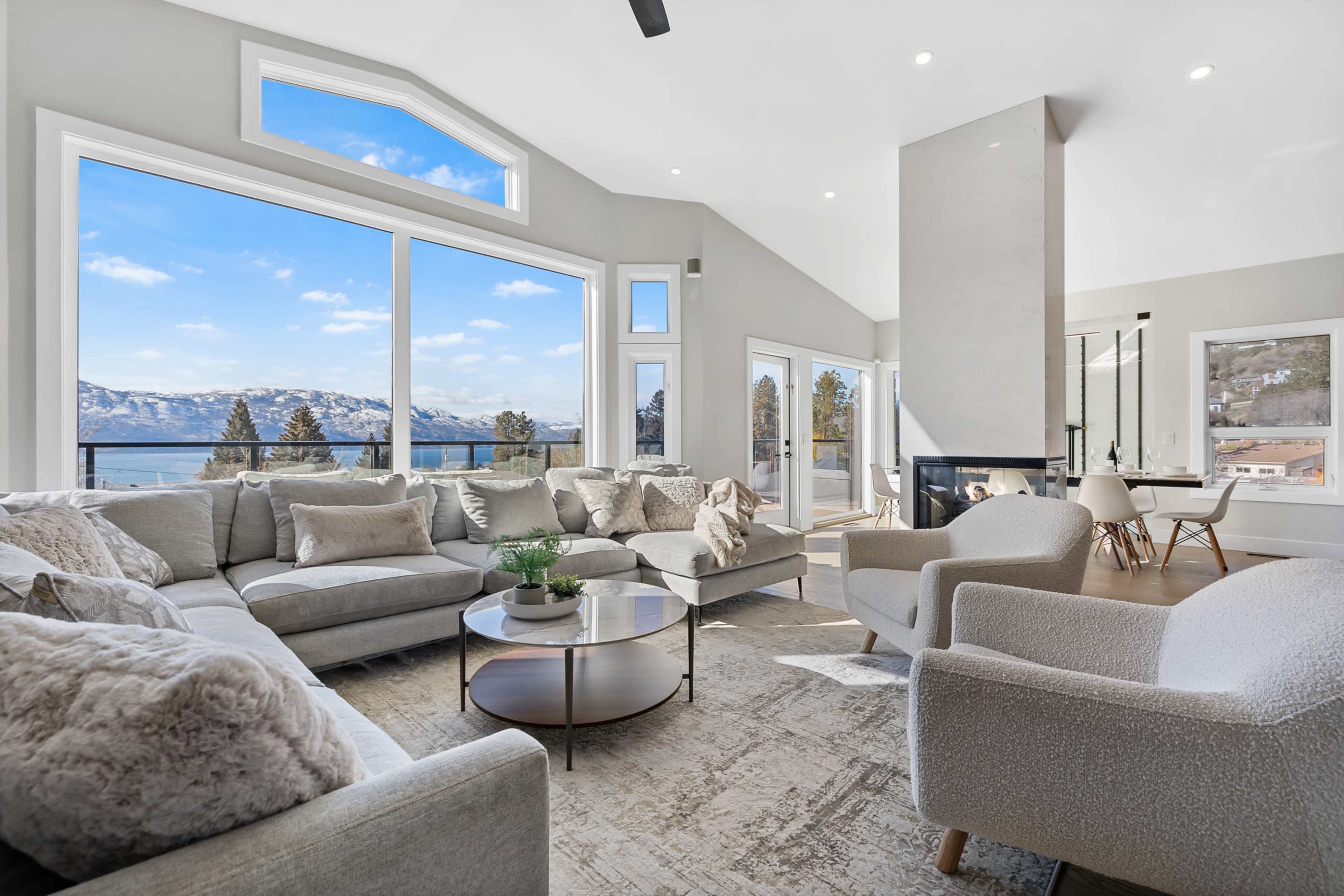 A bright living room with large windows overlooking a lake and mountains, featuring a light gray sectional sofa, a round coffee table, and modern decor.