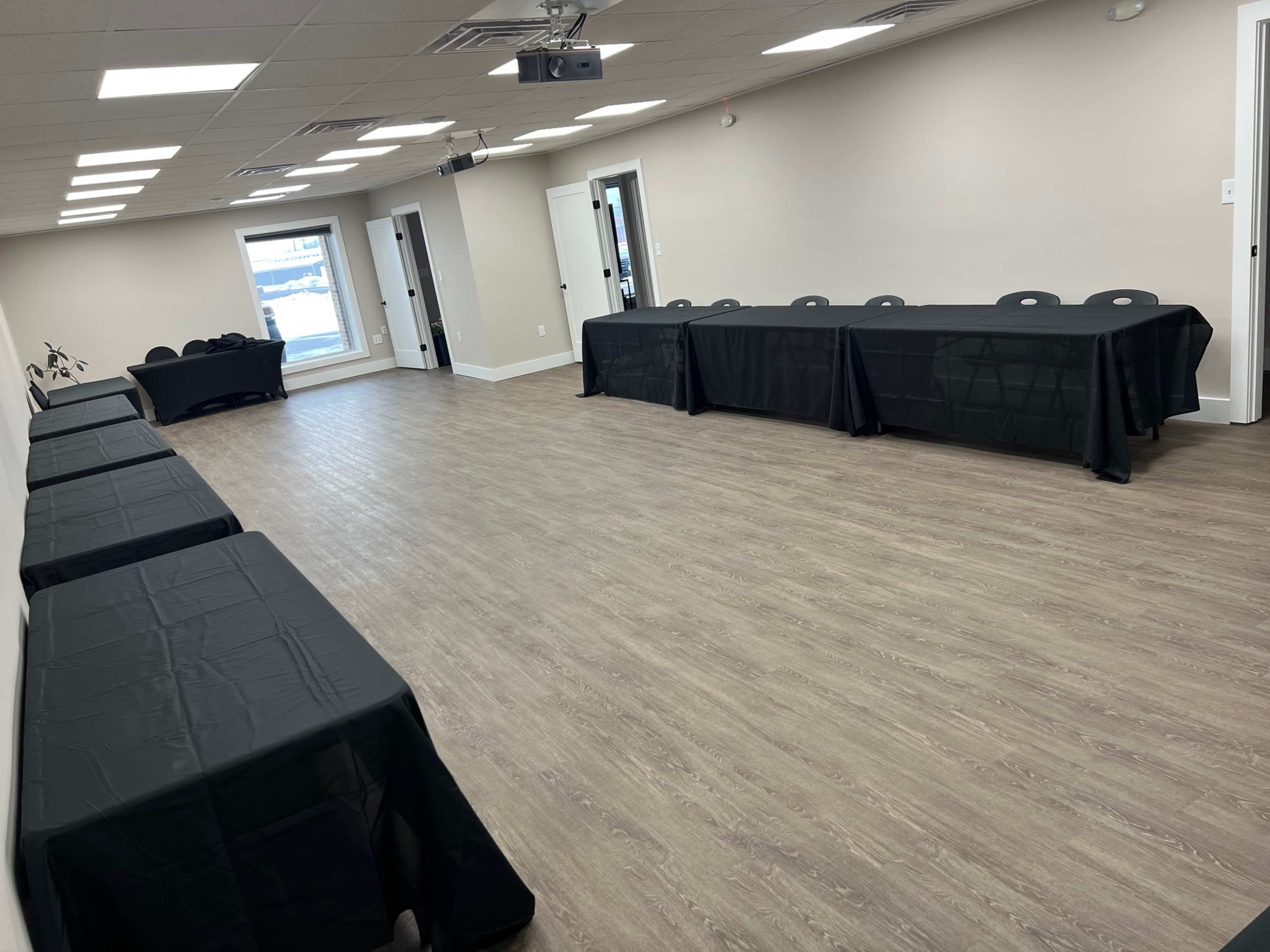 The image shows a spacious room with several black-covered tables arranged in a row against the walls and an empty area in the center, illuminated by overhead lighting.