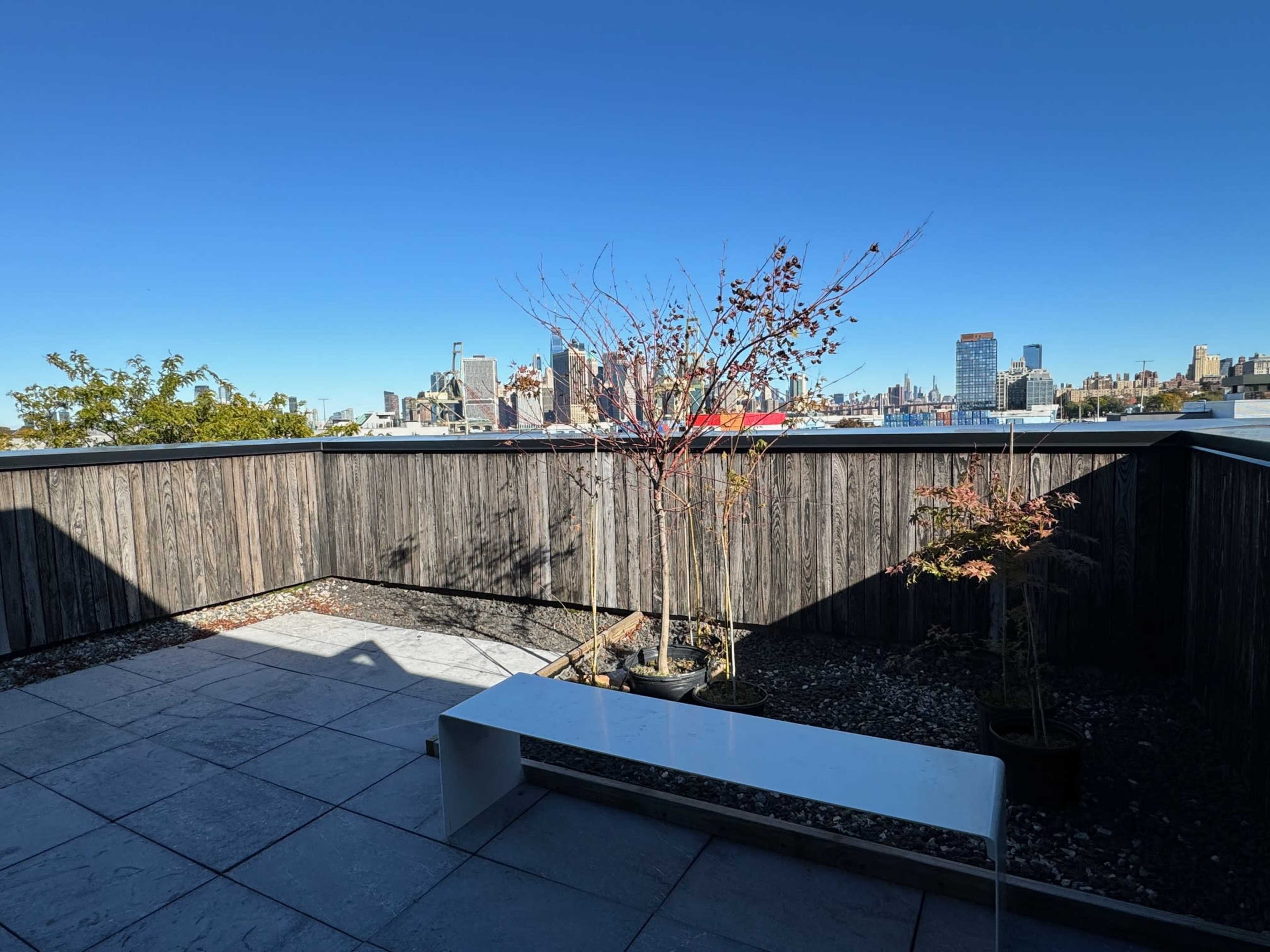 A rooftop terrace features a white bench and small trees, overlooking a city skyline against a clear blue sky.