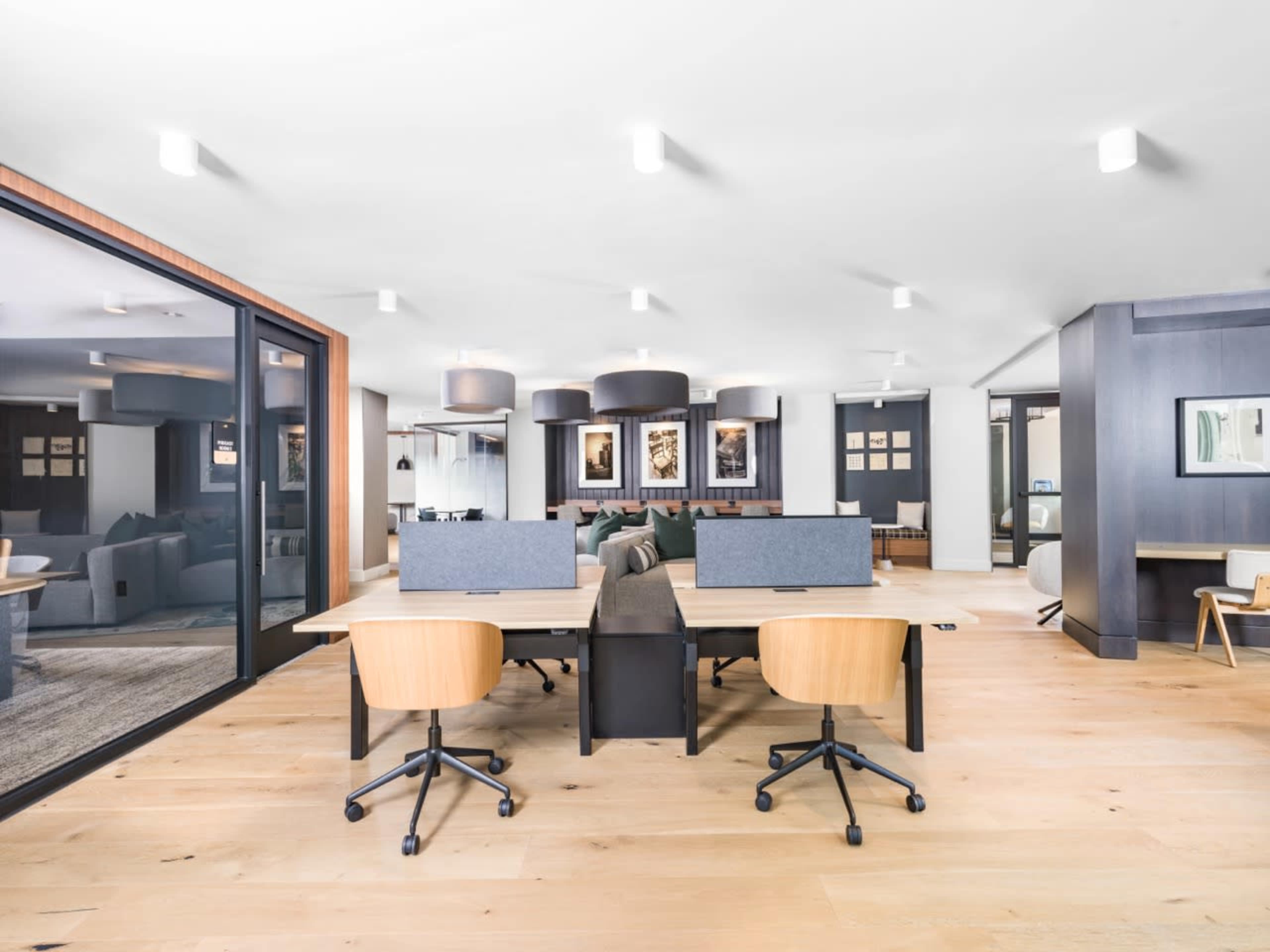 The image shows a modern office space with wooden floors, two desks with chairs, and a seating area in the background.