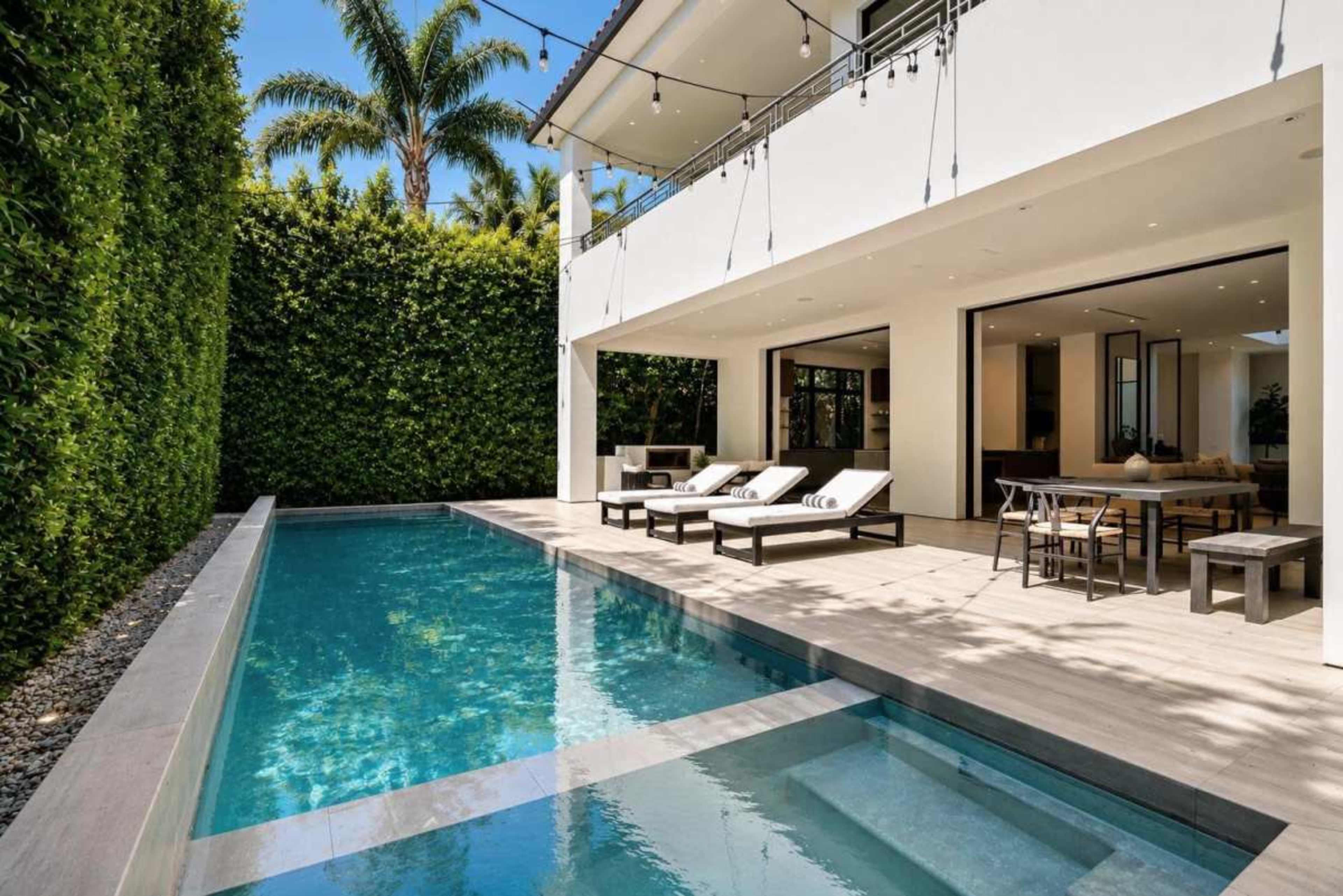 The image shows a modern outdoor area featuring a swimming pool bordered by lounge chairs and a dining table, surrounded by lush greenery and a two-story white building.
