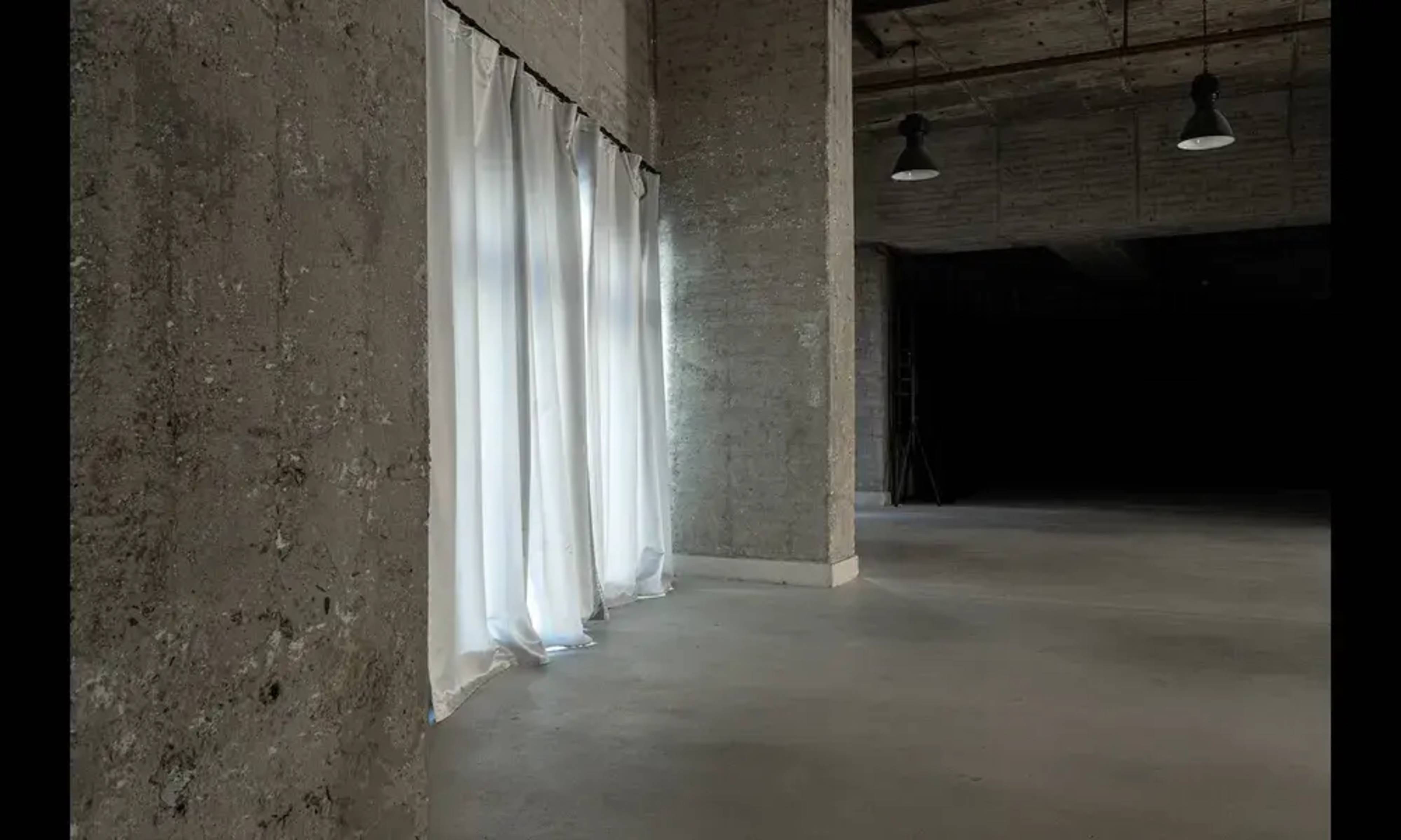 The image shows a spacious, minimalist interior with concrete walls, white curtains at a window, and dim lighting.