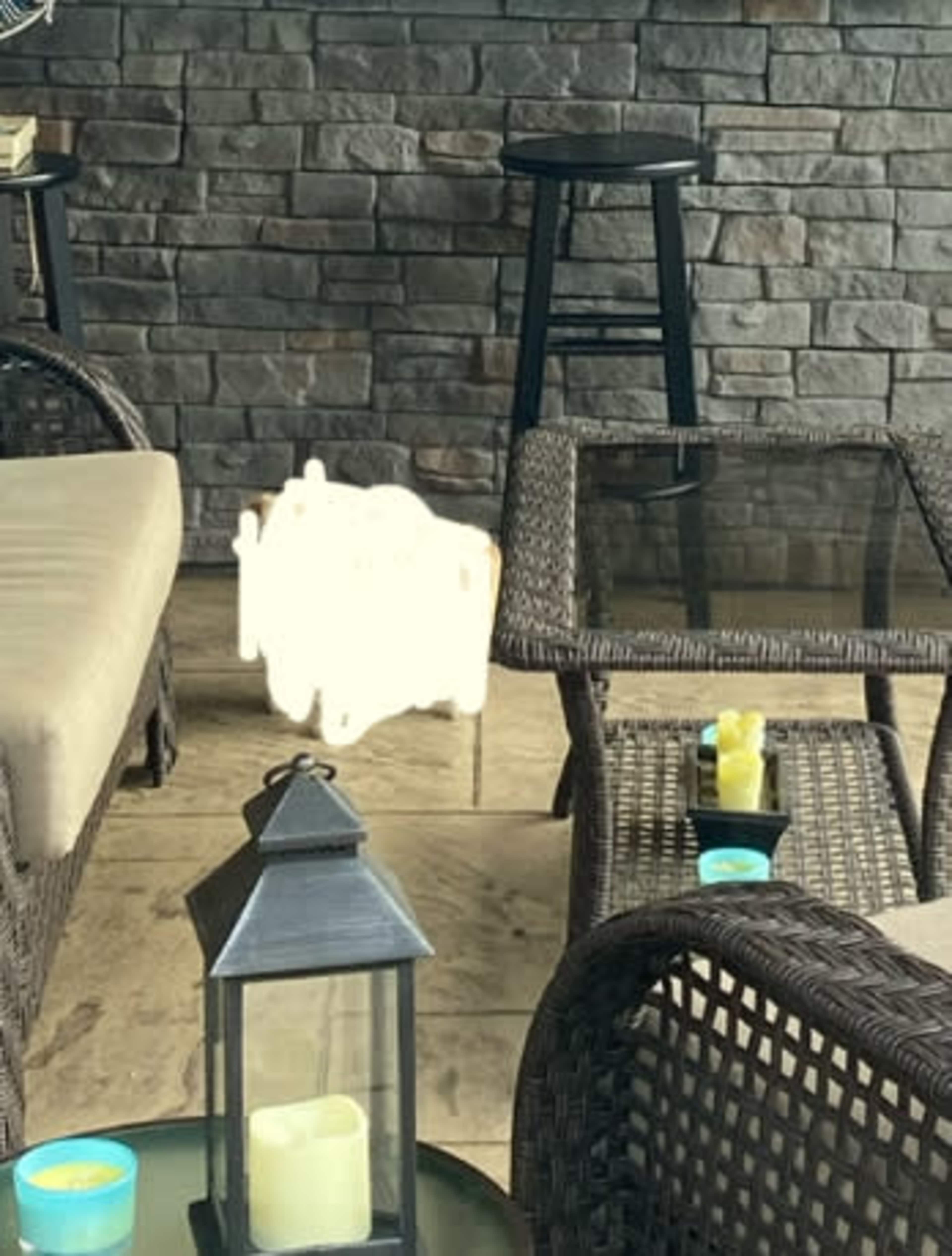 The image shows a cozy seating area with woven furniture, a glass table, and decorative lanterns, set against a stone wall.