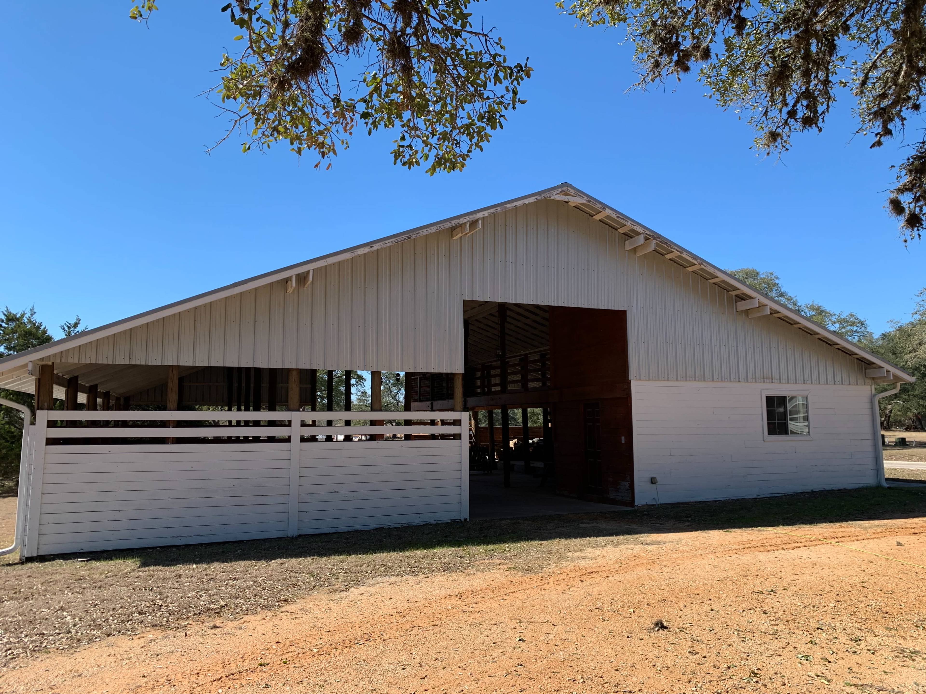A large white barn with a peaked roof sits beside a dirt path under clear blue skies.