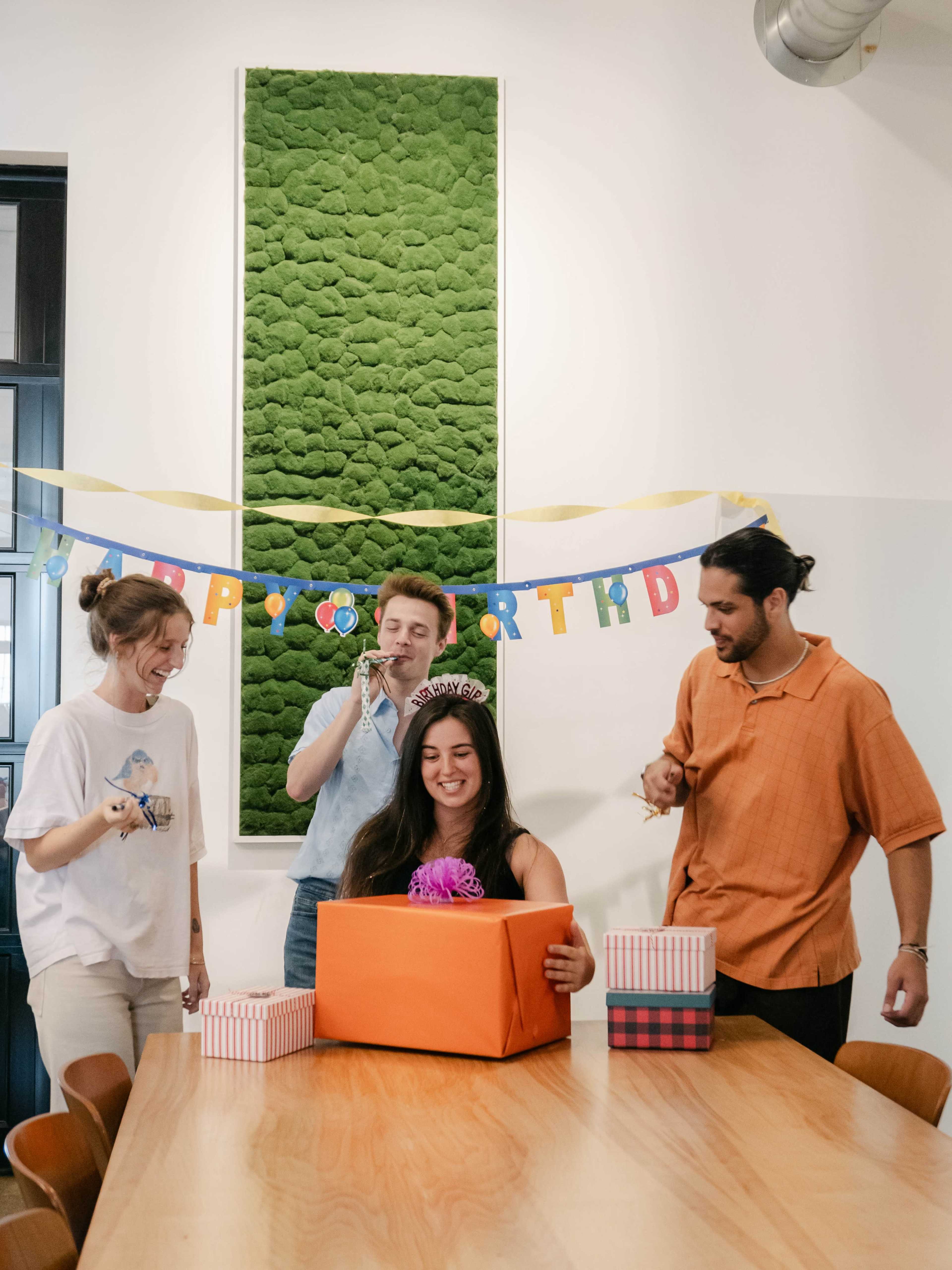 A group of four people gathers around a table in a bright room, celebrating a birthday as one person opens a large orange gift box.
