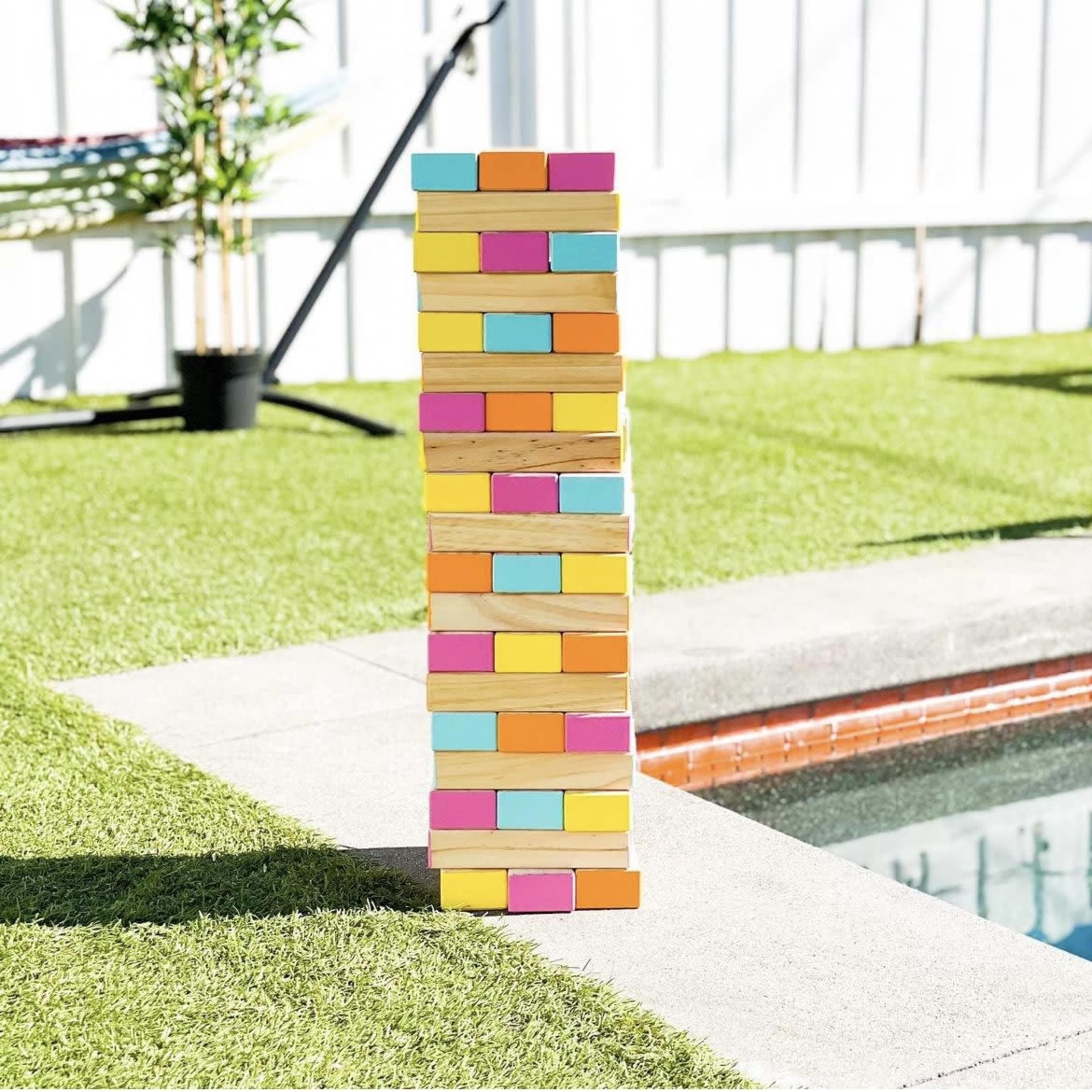 A colorful stack of wooden blocks is balanced on a grassy area near a pool.