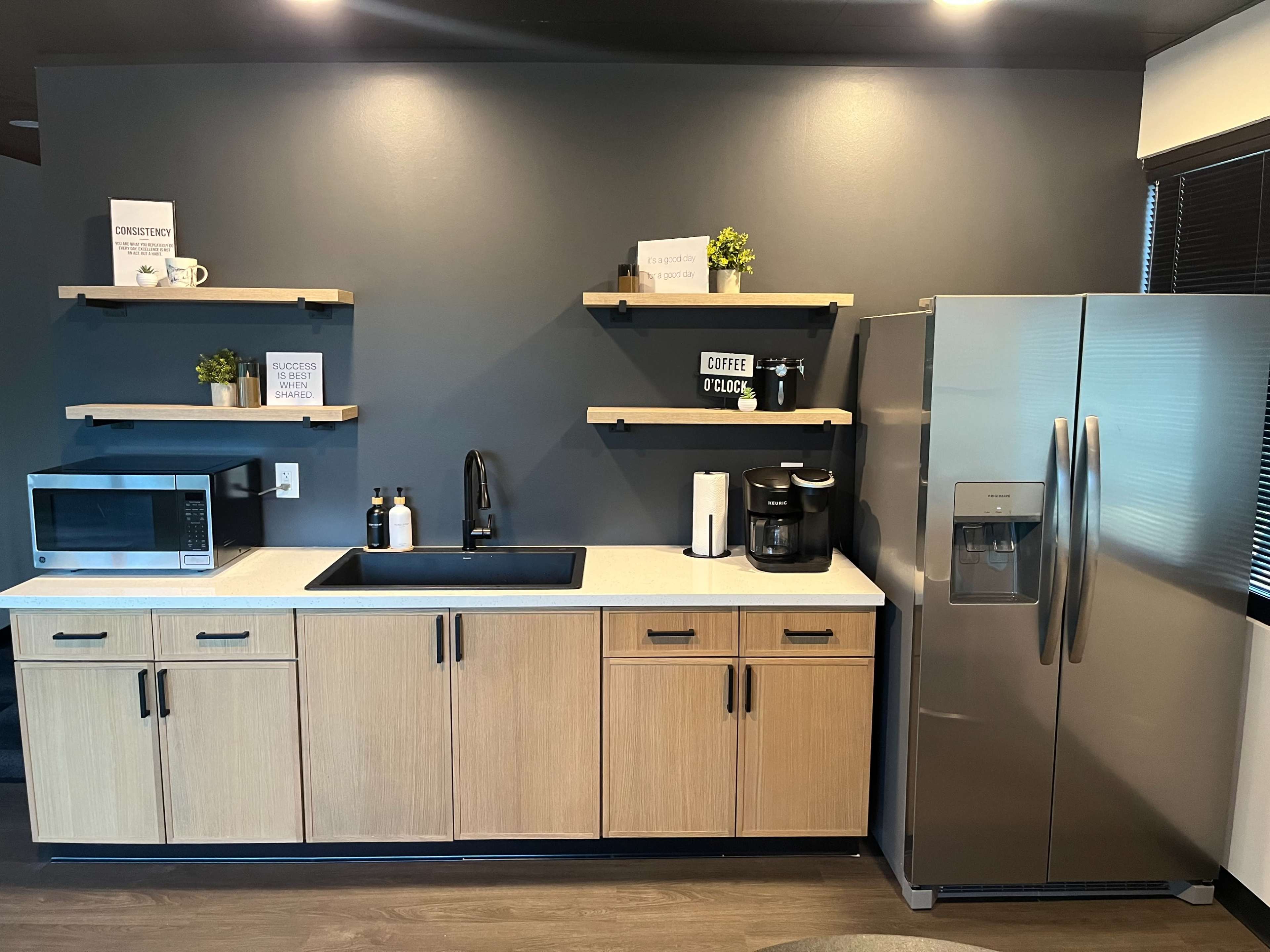 A modern kitchen features wooden cabinetry, a stainless steel refrigerator, a microwave, and floating shelves with decor items.
