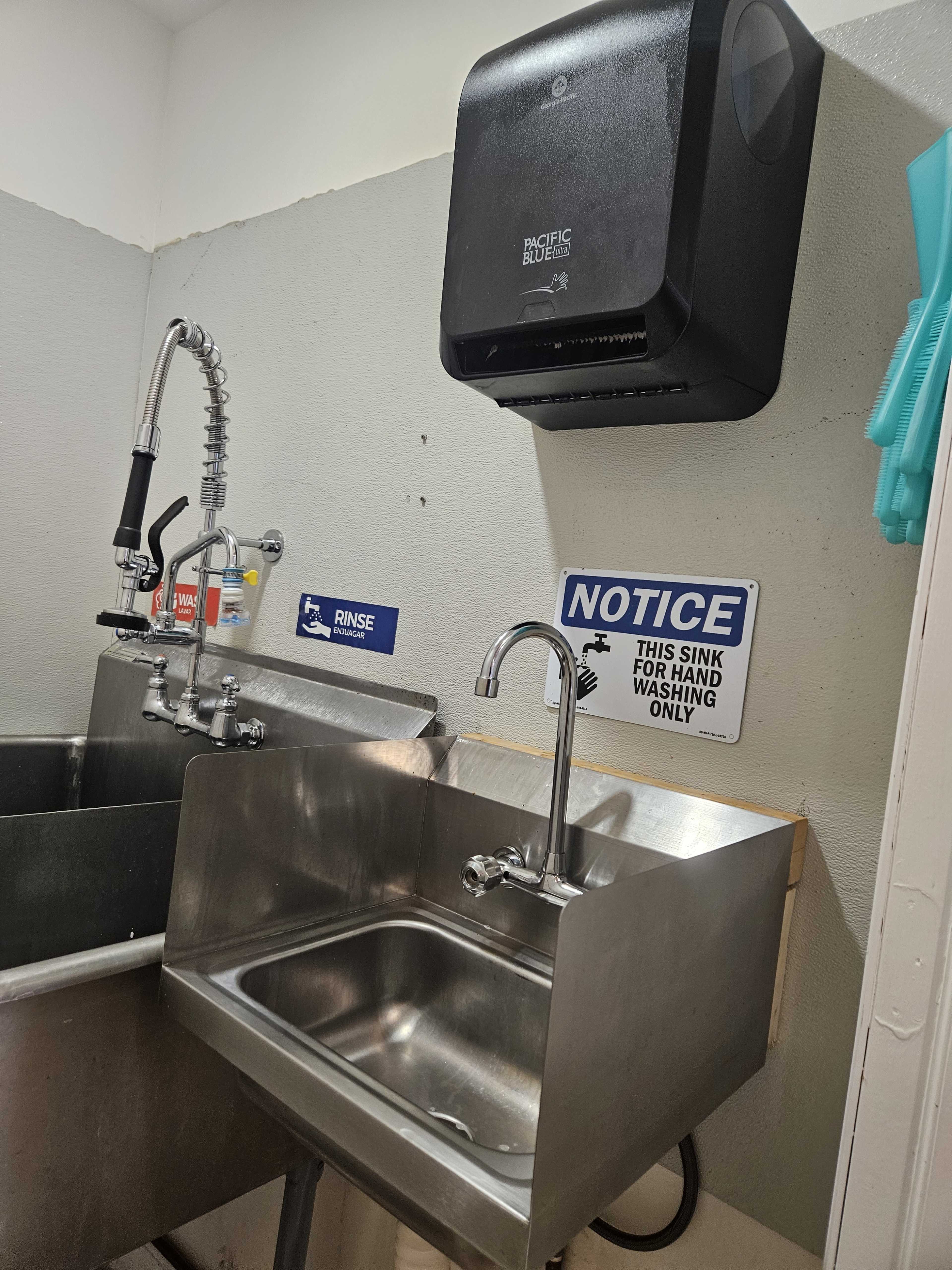 A metallic sink area with a large industrial faucet, a wall-mounted paper towel dispenser above, and signs indicating usage instructions.