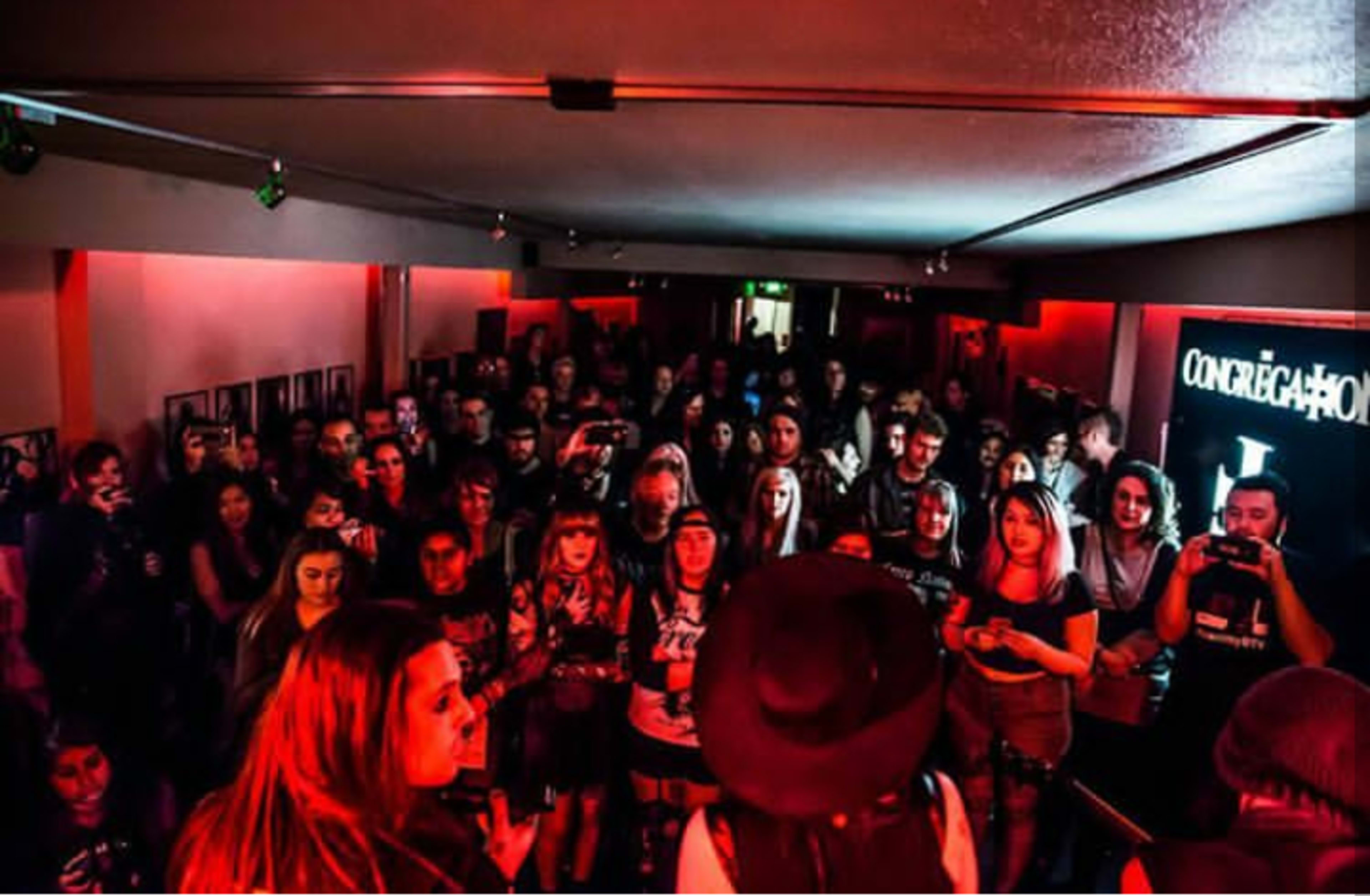 A large crowd stands in a dimly lit venue, attentively watching a performance while illuminated by red lights.