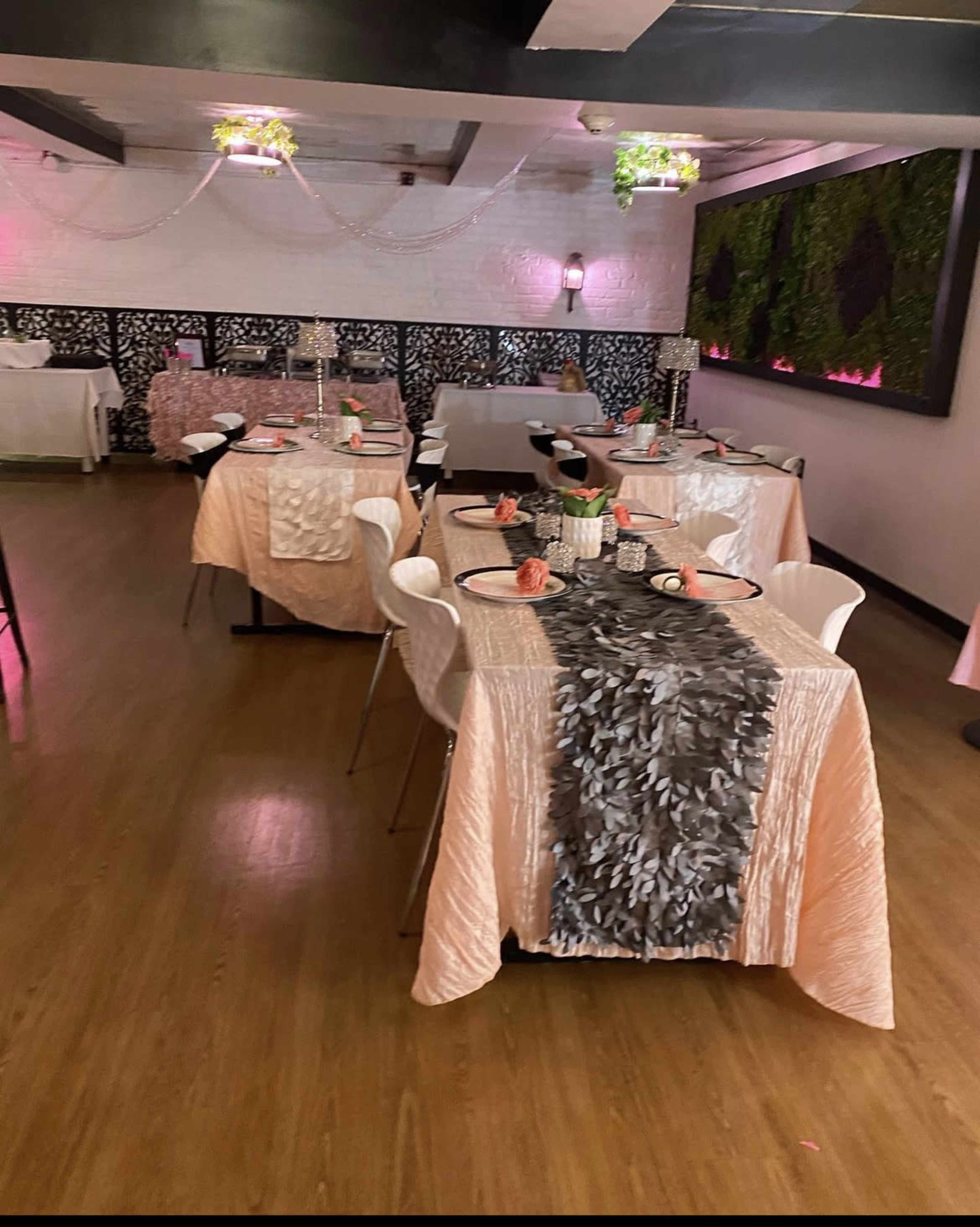 A restaurant interior with several elegantly set tables featuring light pink and gray tablecloths, decorative centerpieces, and a designed wall with greenery.