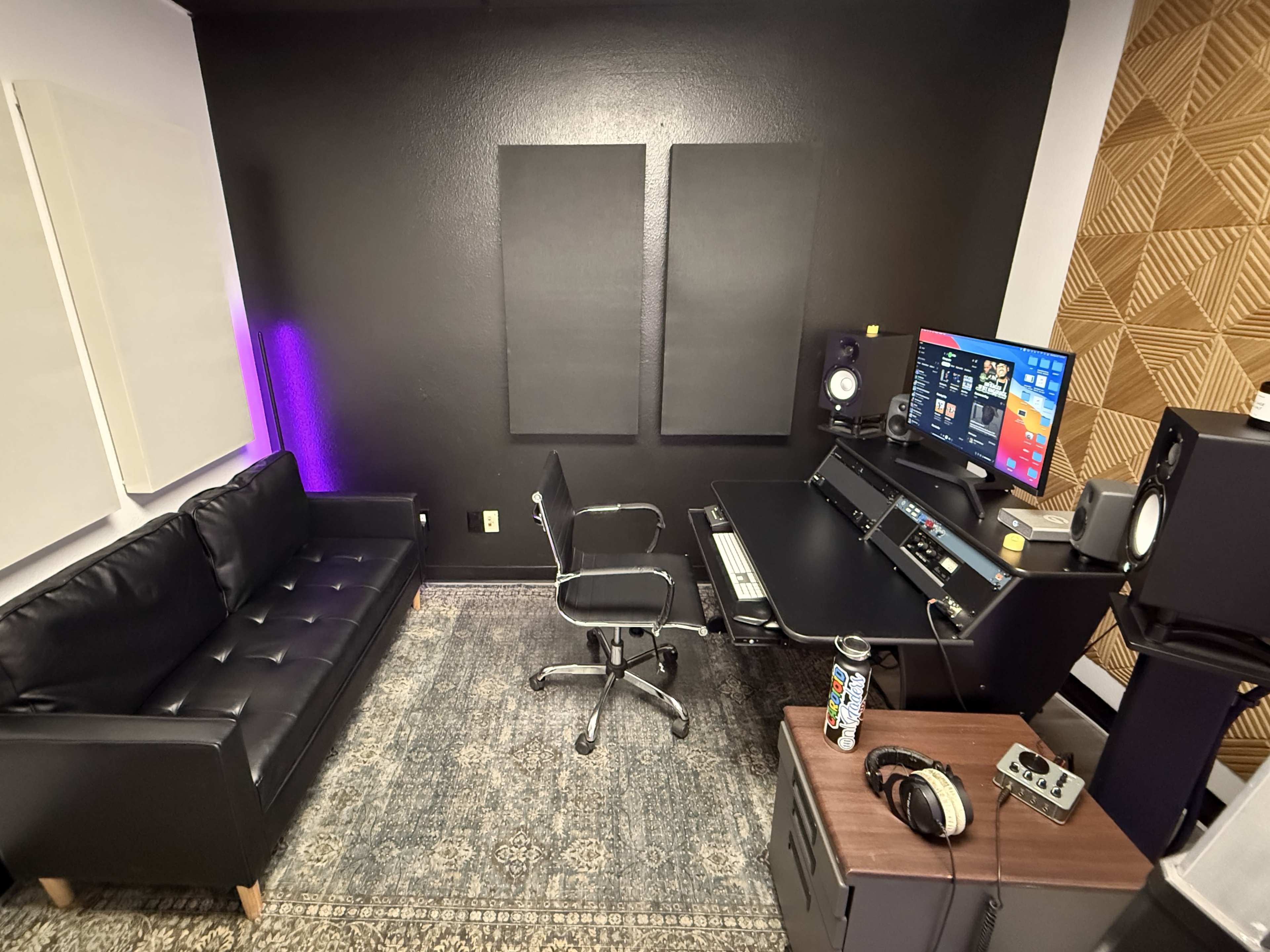 The image shows a recording studio with a black couch, a desk equipped with a computer and sound equipment, and acoustic panels on the walls.