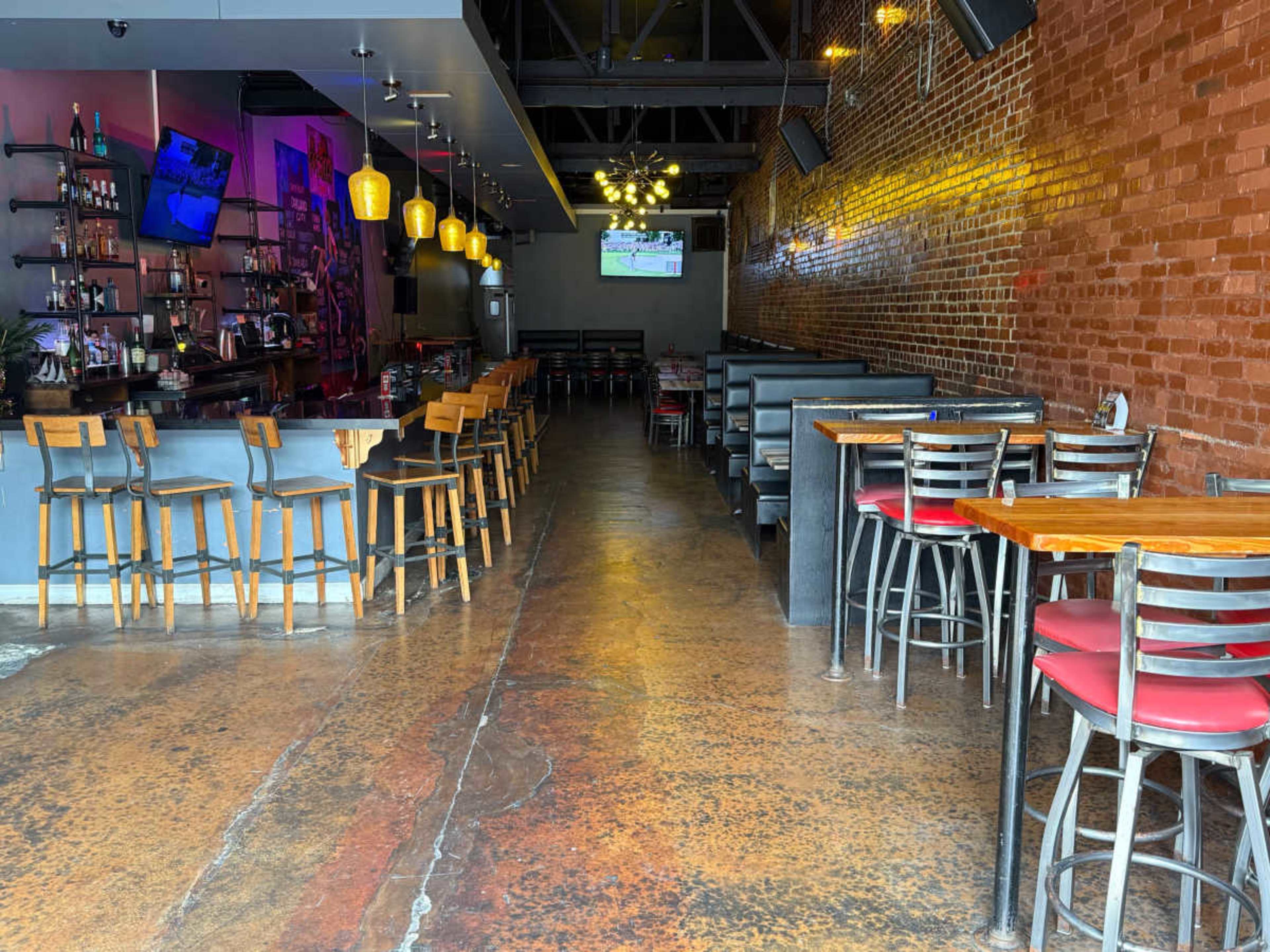 The image shows a bar and dining area with wooden tables and black booths, featuring a long counter with high stools and illuminated pendant lights above.