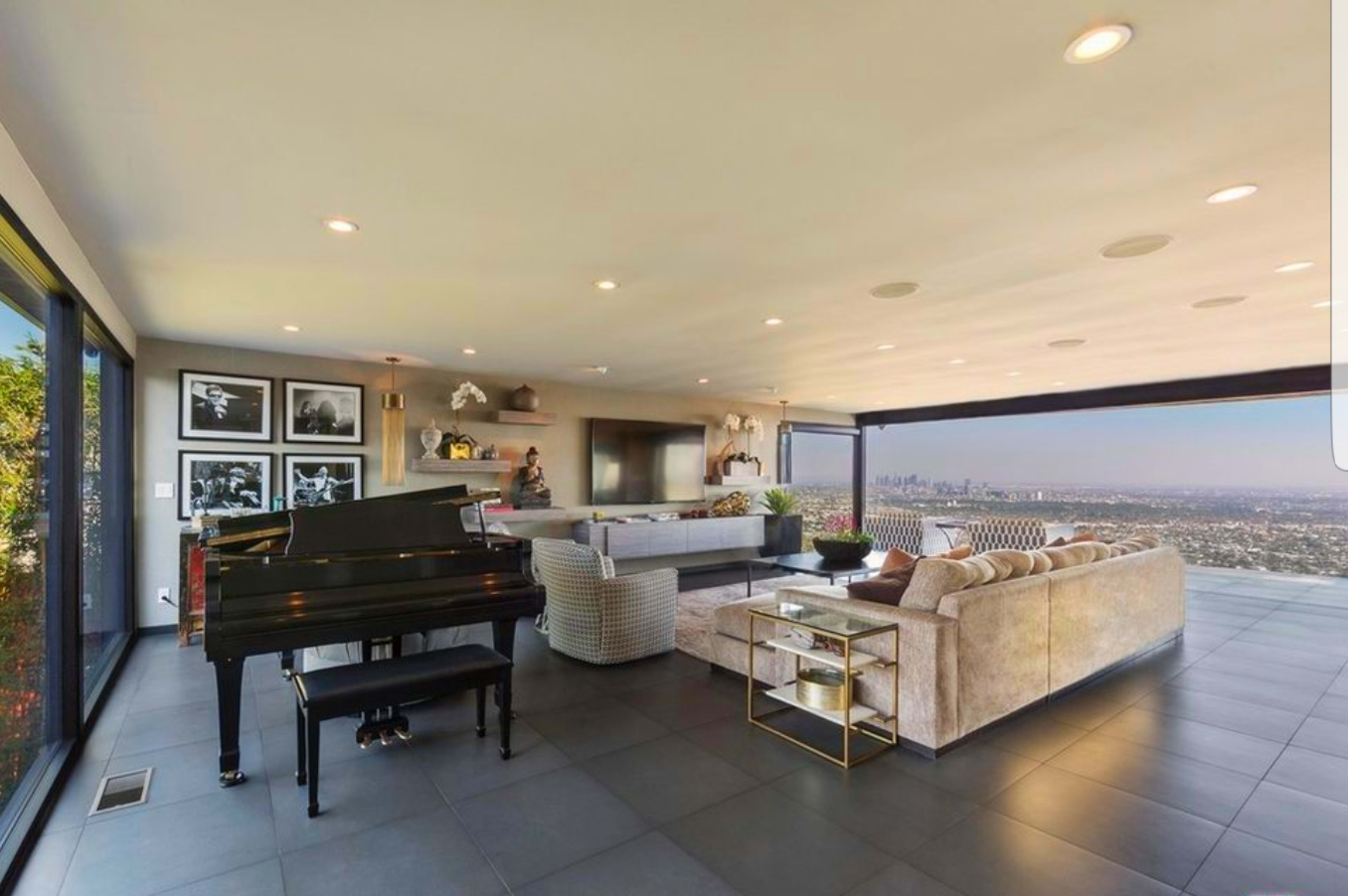 A modern living room with a grand piano, comfortable seating, large windows offering a city view, and minimalist decor.