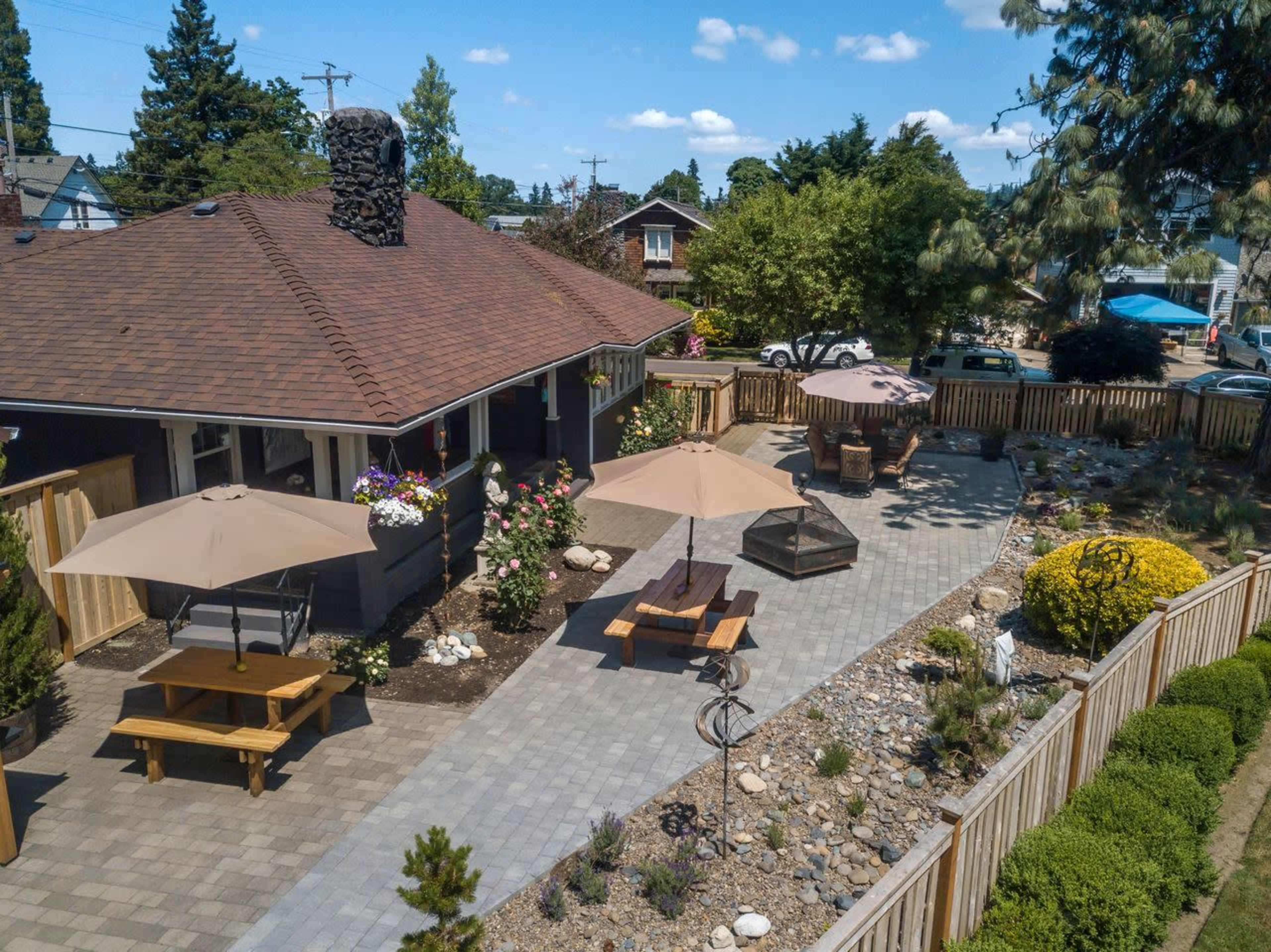 An aerial view shows a backyard patio area featuring a stone path, wooden picnic table, and umbrellas, surrounded by landscaped gardens and a wooden fence.