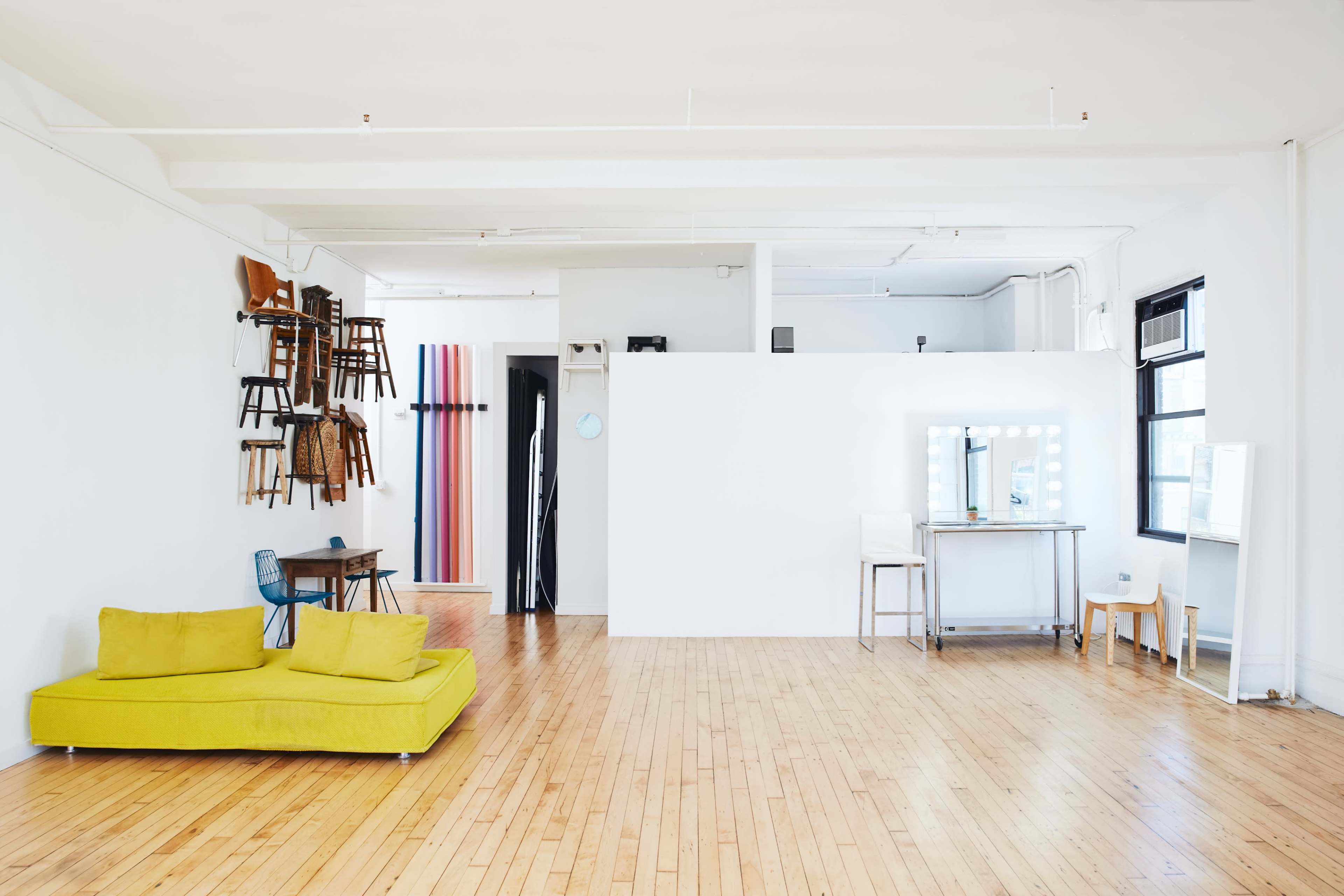 The image shows a spacious, minimalist room with a bright yellow couch, wooden flooring, and a mix of chairs displayed on the wall.
