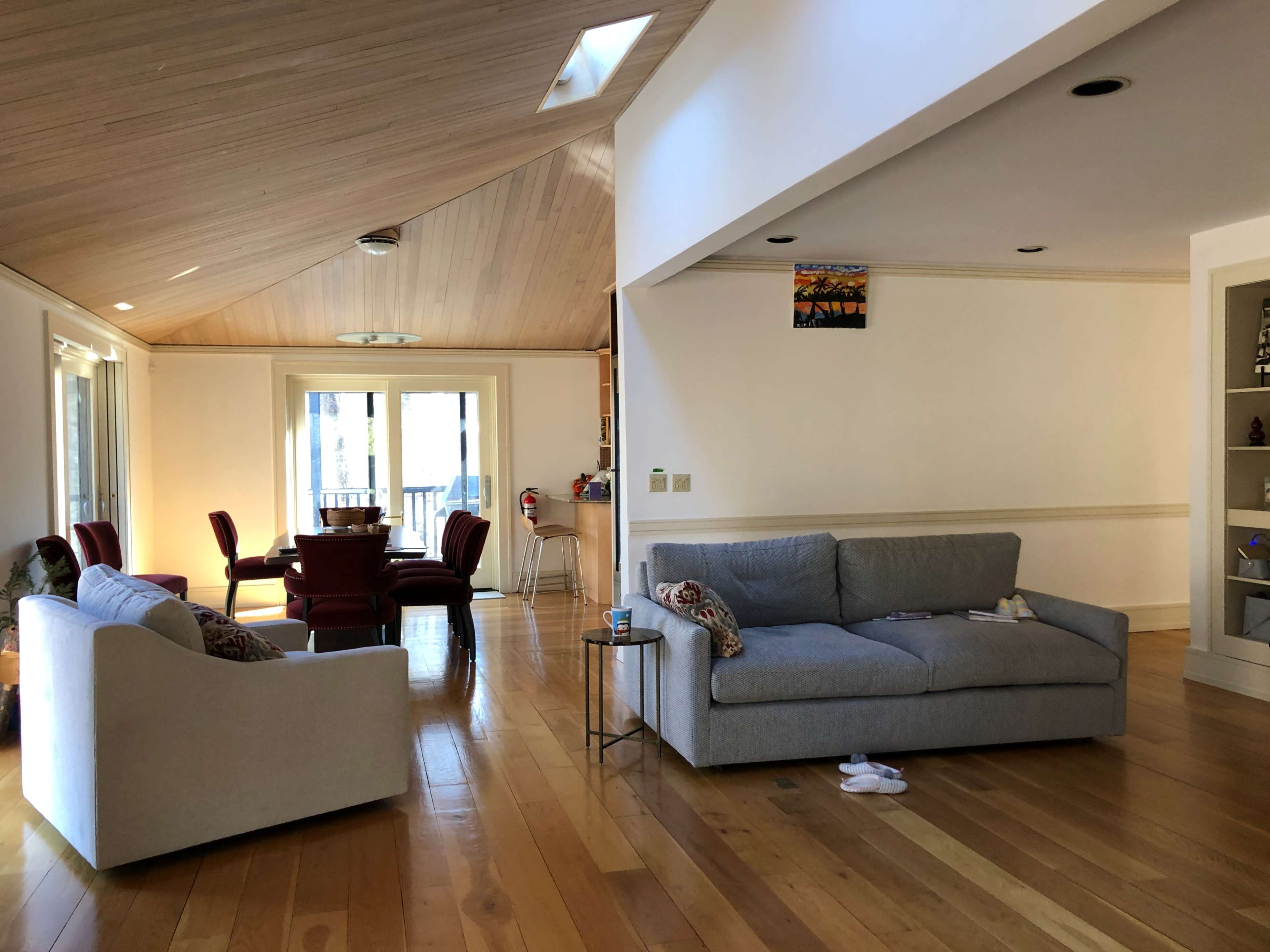 A modern living room with light wooden flooring, a gray sofa, and a dining area featuring a table and chairs in the background.