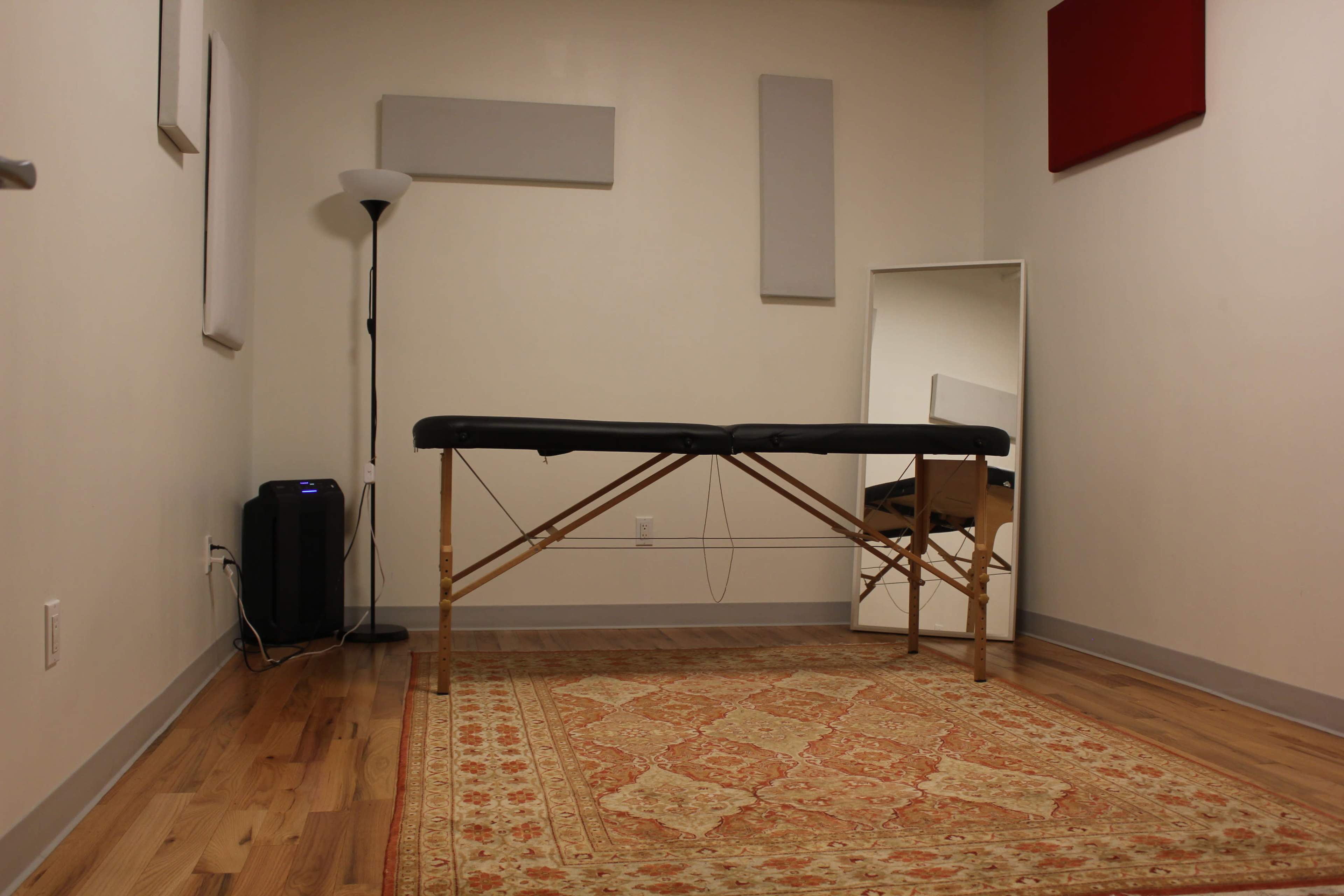 A sparsely furnished room with a massage table, a chair, a mirror, a lamp, and sound-absorbing panels on the walls.