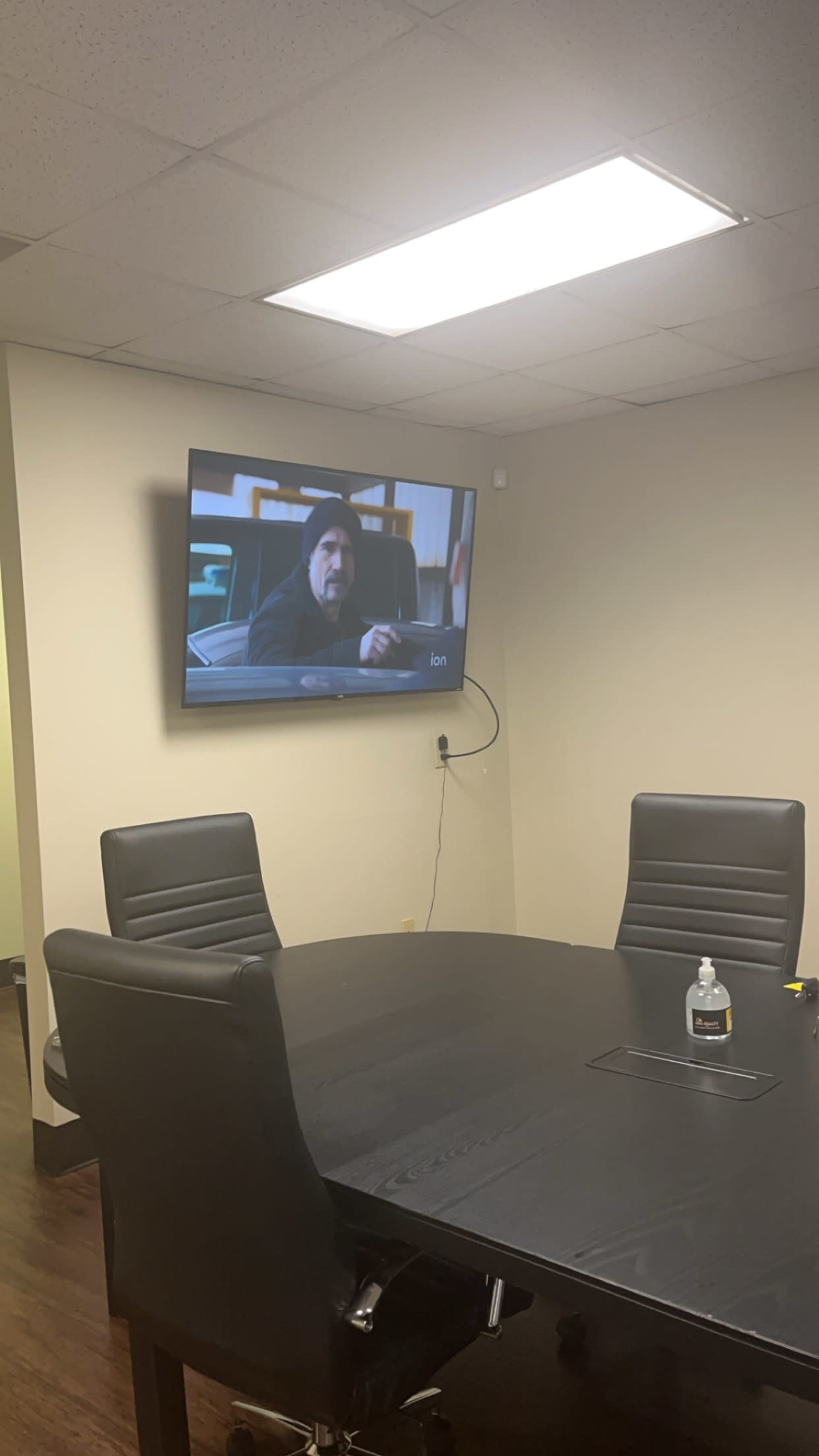 A television displaying a scene from a show is mounted on the wall of a conference room with a round table and office chairs.