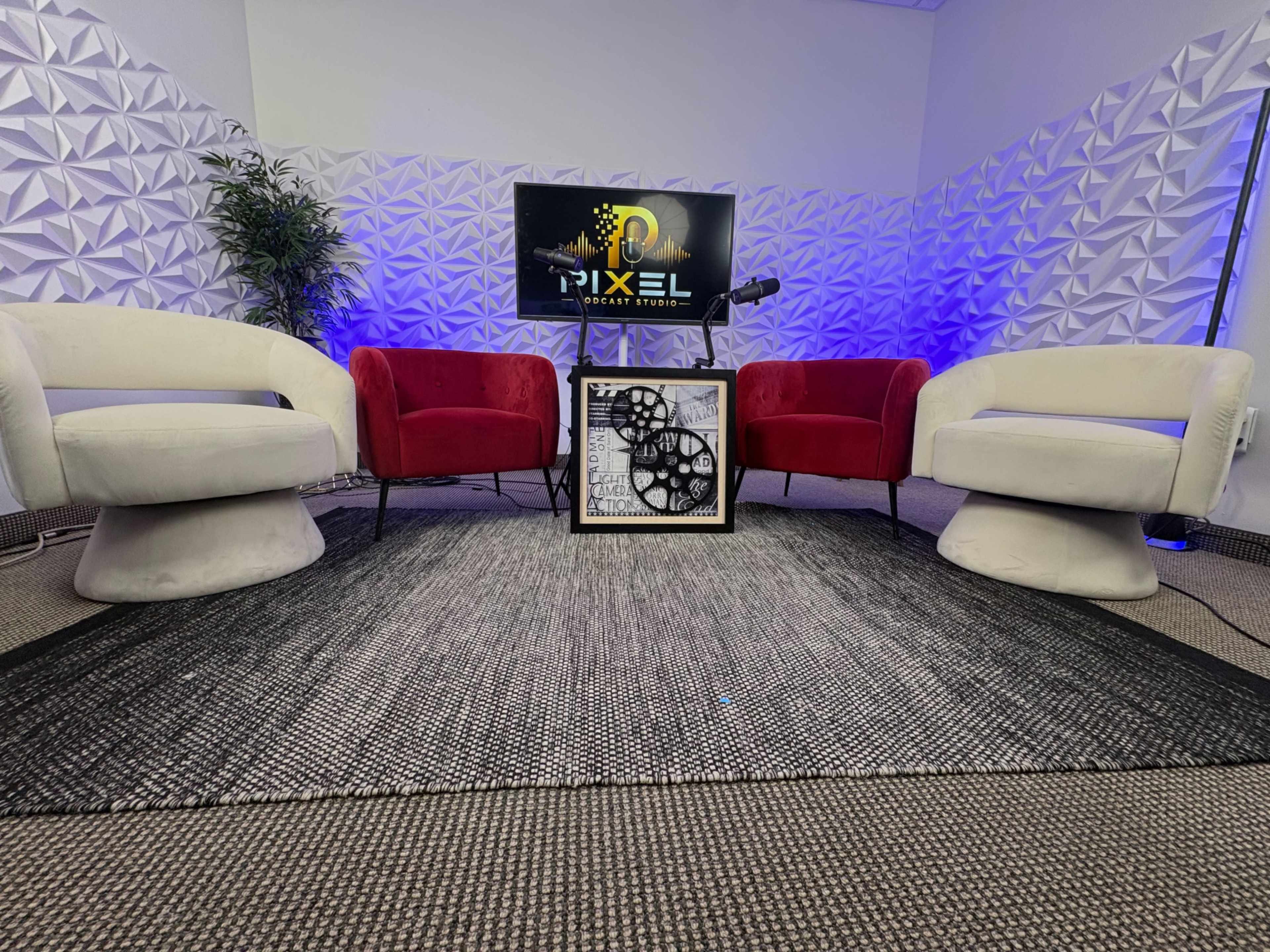 The image shows a small studio setup with two red armchairs and two white swivel chairs arranged around a black and white patterned table, with a backdrop featuring a logo on a screen and decorative wall panels.