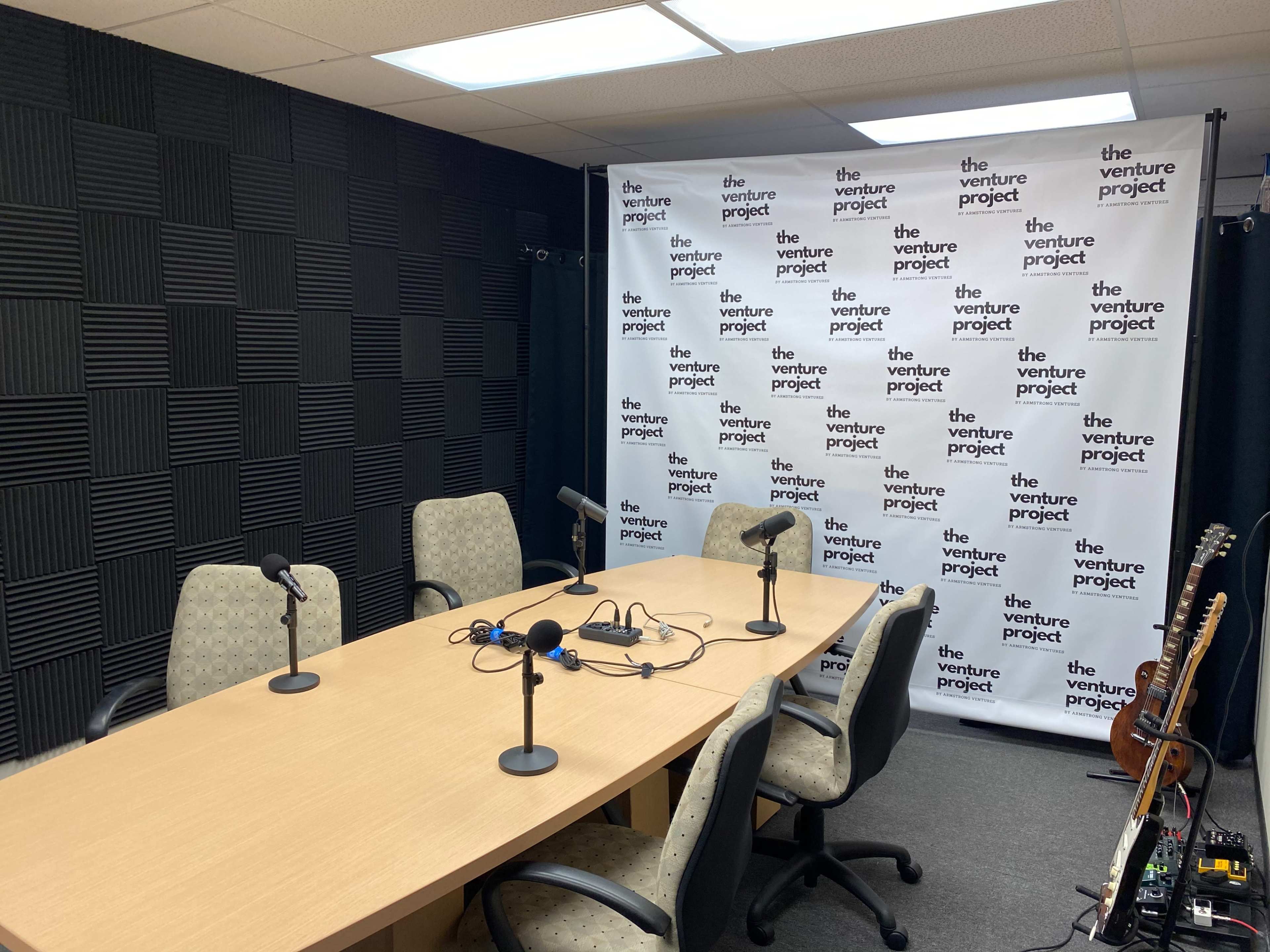 A recording studio with a wooden table, three chairs, and microphones set up in front of a backdrop featuring the words "the venture project."