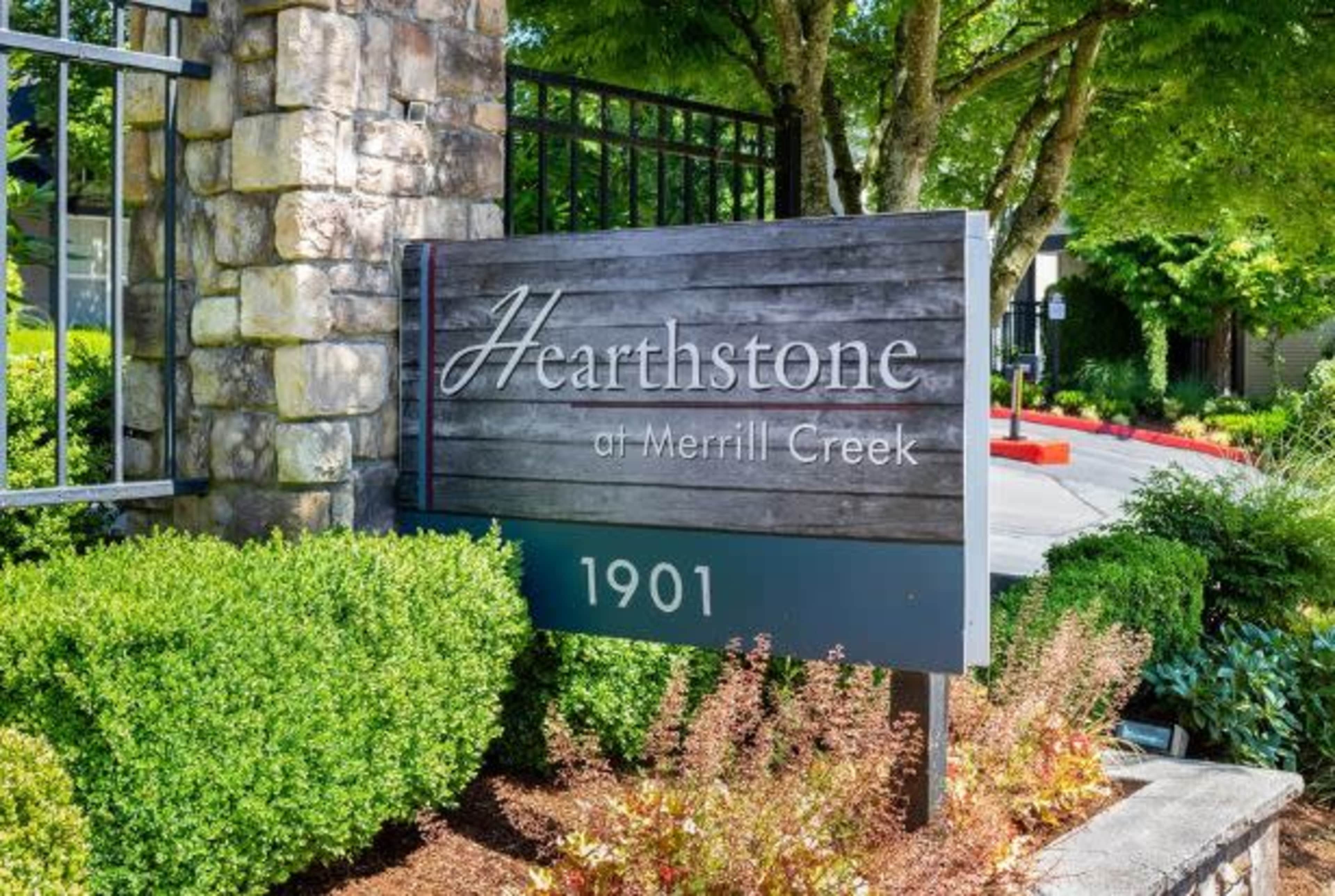 The image shows a sign for "Hearthstone at Merrill Creek," located at 1901, surrounded by neatly trimmed shrubs and vibrant foliage.